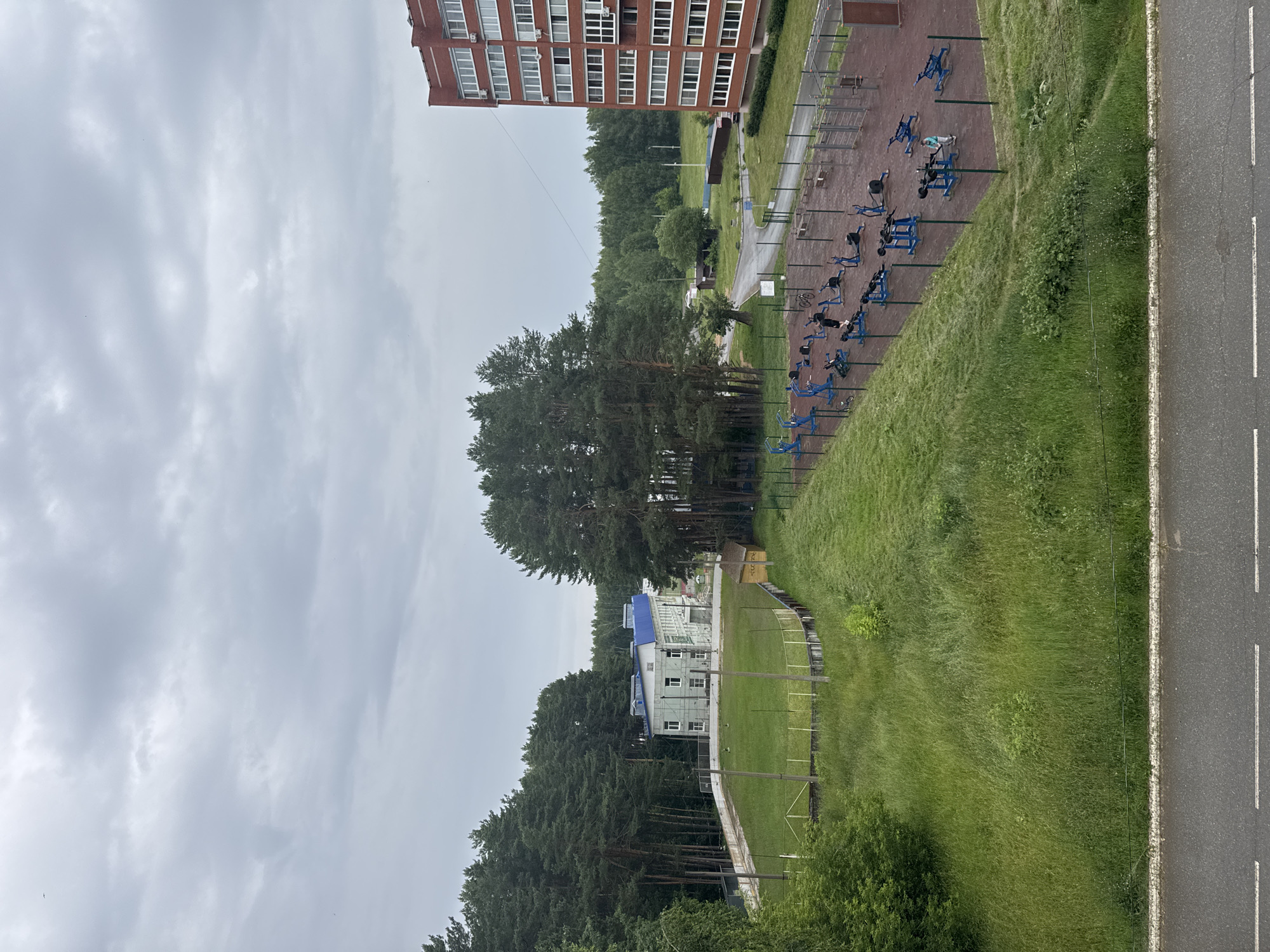 The image depicts an outdoor fitness area situated in a residential setting, likely in Chaikovsky, Russia. In the foreground, a row of blue outdoor exercise machines is visible on a red rubberized surface, with a few individuals using the equipment. Adjacent to the fitness area is a steep, grassy embankment that slopes down towards a road.
Further in the background, a multi-story brick building stands tall, its windows reflecting the overcast sky. Beyond this, a cluster of tall pine trees forms a dense forest. A smaller, white building with a blue roof is nestled amongst the trees, suggesting a park or recreational facility. The overall scene is one of an urban environment integrated with nature, offering recreational opportunities for residents.
The weather appears to be cloudy, indicated by the gray, diffused light and the texture of the clouds in the sky. The time of day is indeterminate but suggests daylight hours. No visible text is present in the image. The presence of the fitness equipment and people engaging in exercise implies an ongoing recreational activity within this community space.