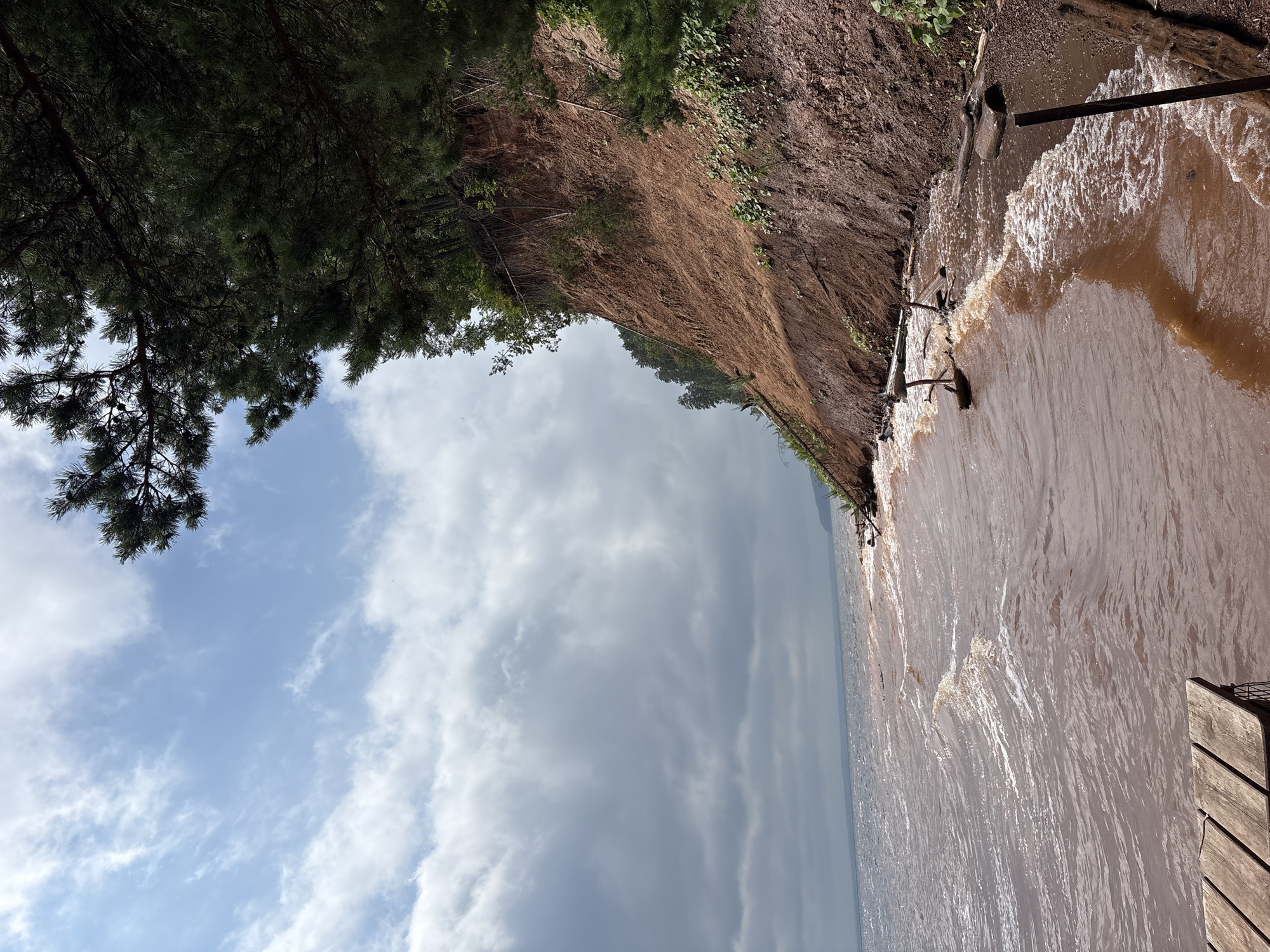 This is a landscape photograph of a rocky shoreline with reddish-brown soil. The water is choppy and appears to be a muddy brown color. A steep, eroded cliff faces the water, covered in sparse green vegetation and trees. The sky is partly cloudy with patches of blue. In the foreground, on the left side, a portion of a wooden dock is visible. The image appears to be taken from a slightly elevated perspective, looking out over the water and along the coastline. The location is specified as Костоваты, Russia.