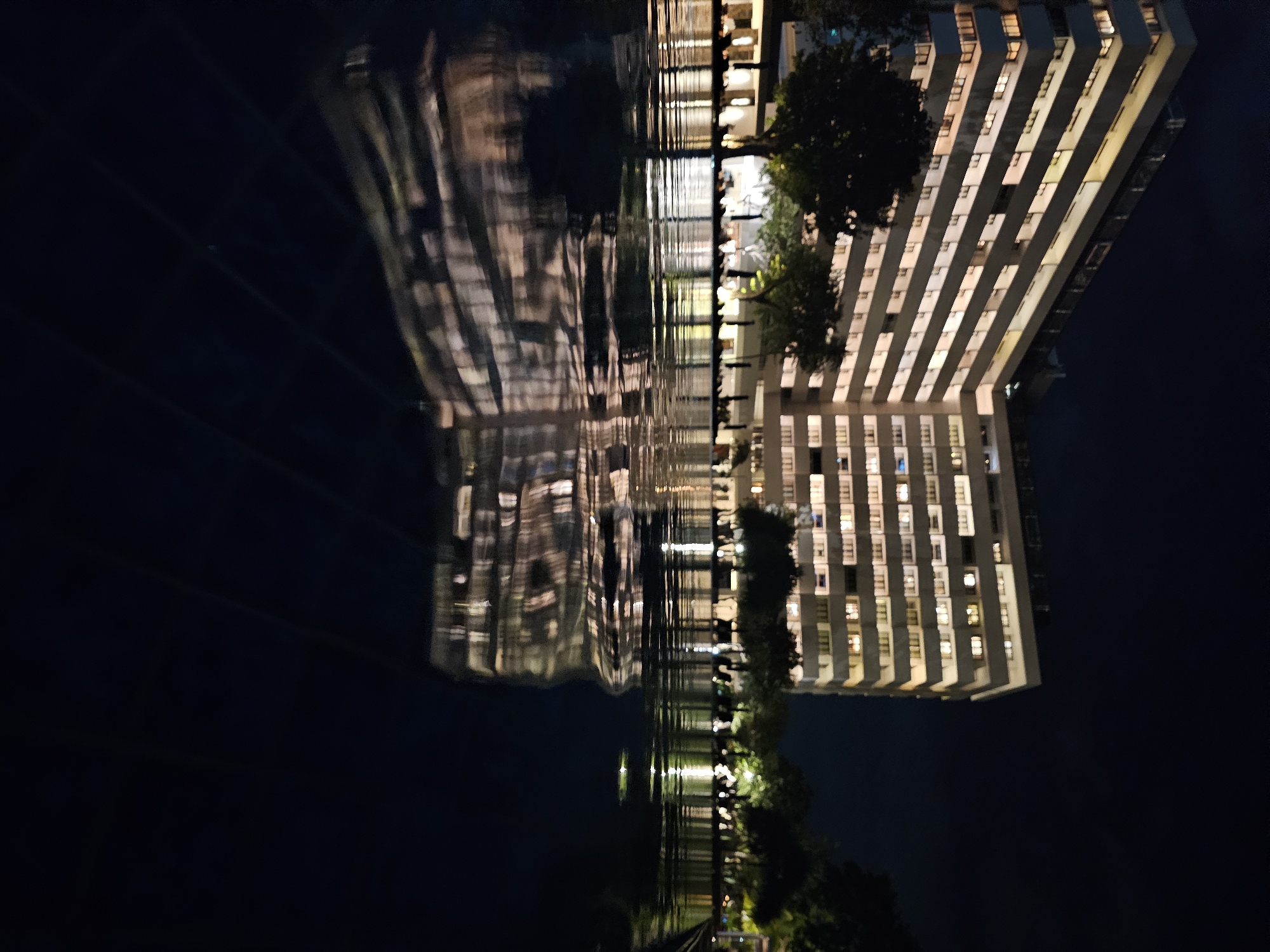 An exterior night shot captures a modern, multi-story building with illuminated windows, reflecting on a still body of water in the foreground. The building appears to be a hotel or residential complex. The architecture is characterized by horizontal lines and balconies, with warm light emanating from many of the windows, suggesting occupancy. Several trees with dark foliage are visible in front of the building, adding a natural element to the urban landscape. The water's surface is smooth, creating a clear reflection of the building and surrounding lights. The overall scene is serene and modern, with the lighting creating a warm and inviting ambiance against the dark night sky. The location is the city of Denpasar, Indonesia.