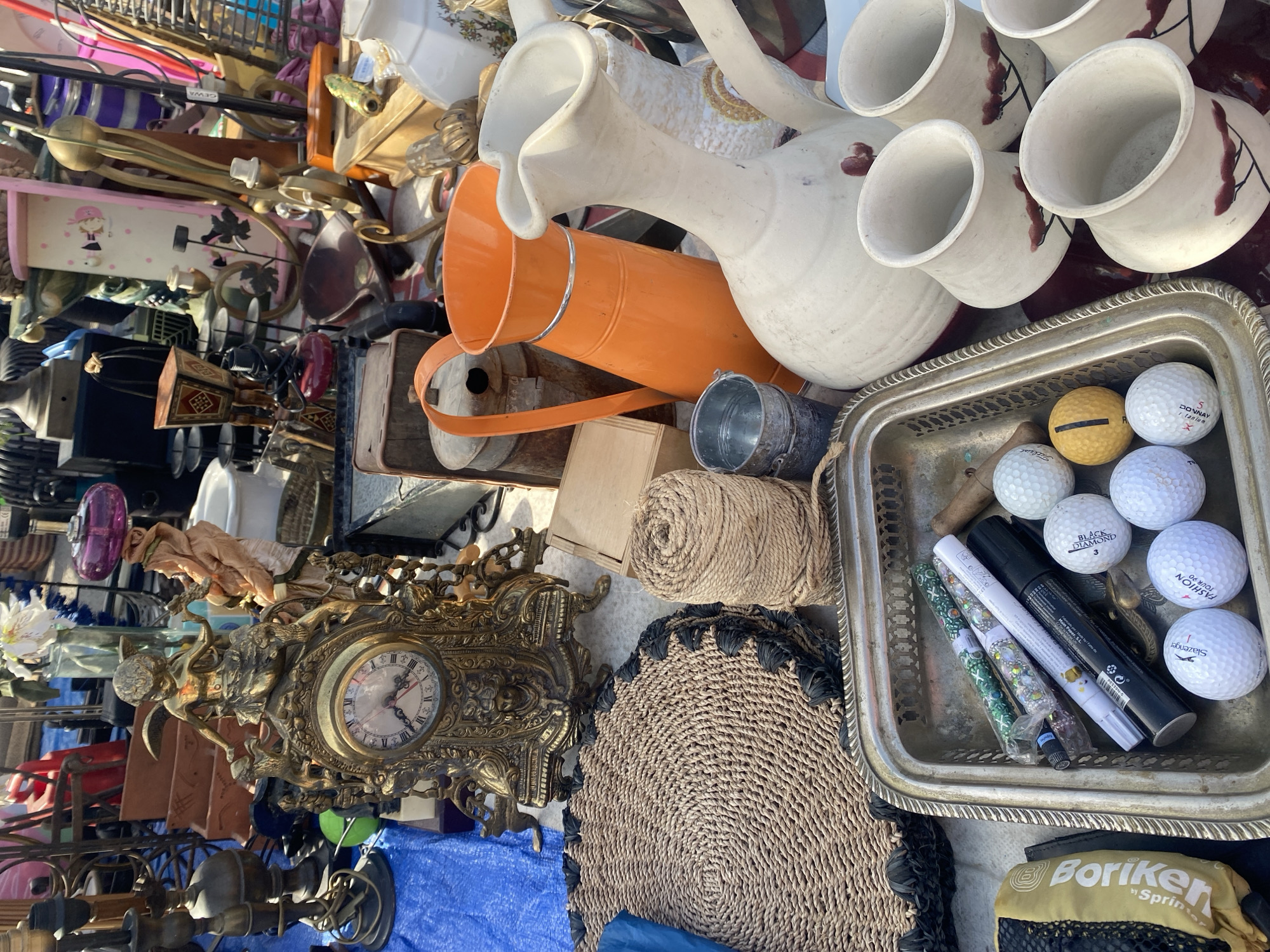 This is a close-up, eye-level, outdoor shot of a flea market stall. The stall is laden with a variety of vintage and used items, including decorative clocks, pitchers, vases, woven mats, golf balls, and writing implements. The overall color palette is earthy and muted, with pops of bright orange and white from the ceramicware and pitchers. The lighting suggests a bright, sunny day, casting short shadows. The items are displayed on tables and on the ground, creating a sense of abundance and variety. It is implied this market is in Manilva, Spain.