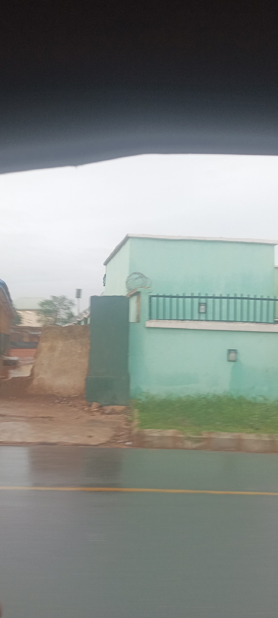 The image depicts a section of a street in what appears to be Umuahia, Nigeria, on an overcast day. The road is wet, and a yellow line marks the lane. On the right, there is a building with a light blue facade and a dark green barred fence. There's grass growing along the base of the fence. Adjacent to the building, there's a section of exposed earth or partially demolished wall, appearing to be made of a reddish-brown material. In the background, other structures are visible, but they are out of focus due to the camera's depth of field and distance. The very top of the frame is obscured by a dark object, likely part of the interior of the vehicle from which the photo was taken. There are no visible people in the shot.