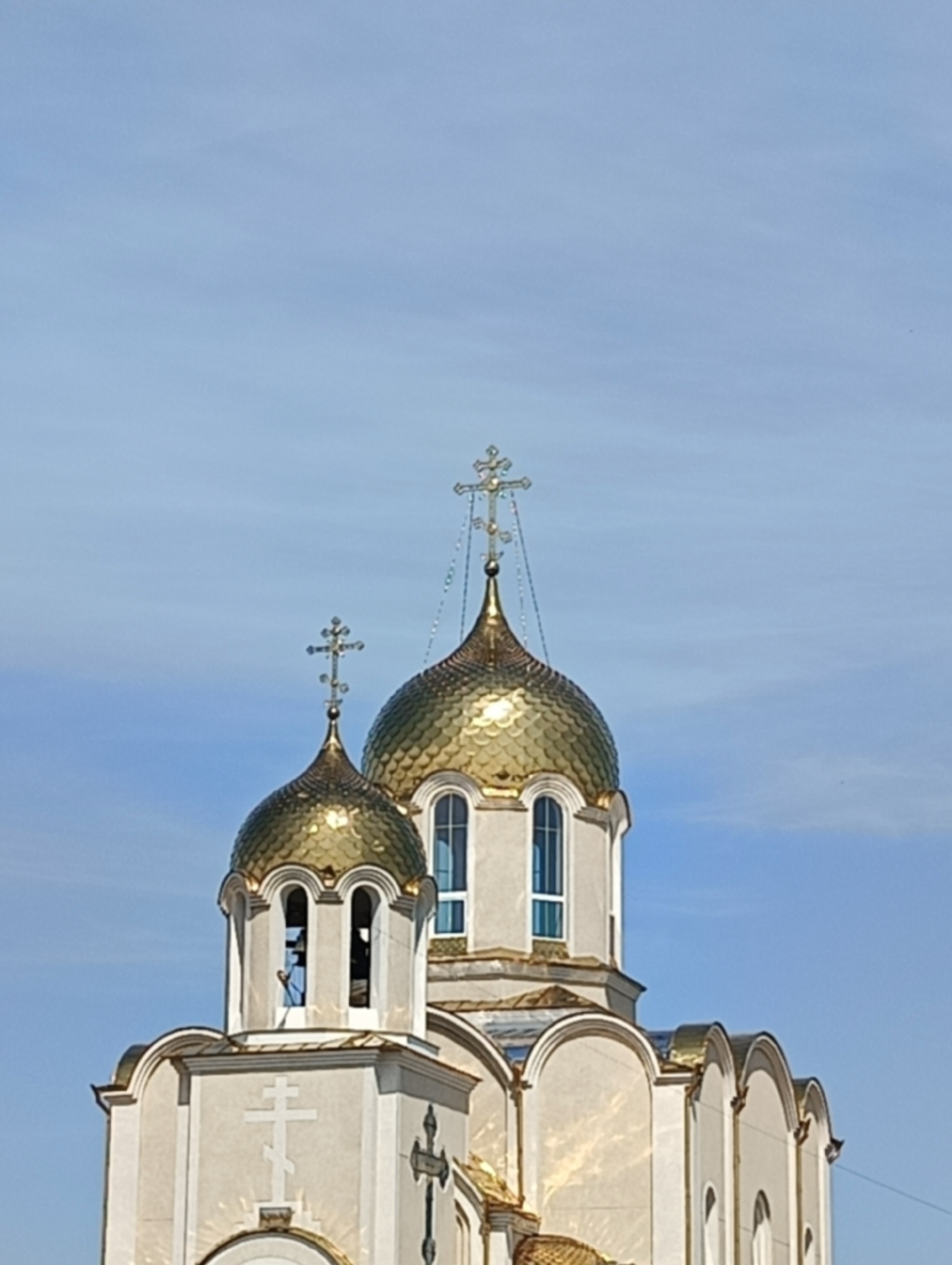 This is an upward-angled photograph of an Eastern Orthodox church located in Maiaky, Ukraine. The central subjects are two prominent golden, scale-patterned onion domes, each crowned with an elaborate Orthodox cross. The larger dome, situated towards the upper right, features a cross with decorative chains descending from its arms. The smaller dome, positioned in the mid-left, rests atop a light-colored belfry-like structure with arched openings, one of which reveals a dark interior. The church building itself is light-colored, likely cream or white, with arched window frames and architectural detailing. Strong sunlight illuminates the scene, creating bright reflections on the golden domes and parts of the building's facade. A flat, white Orthodox cross is affixed to a section of the church wall below the smaller dome, and a darker, metallic cross is partially visible on the right side of the structure. The background is a clear, bright blue sky with faint, wispy clouds. No people, specific actions, or other elements of the surrounding city environment are visible within the frame.