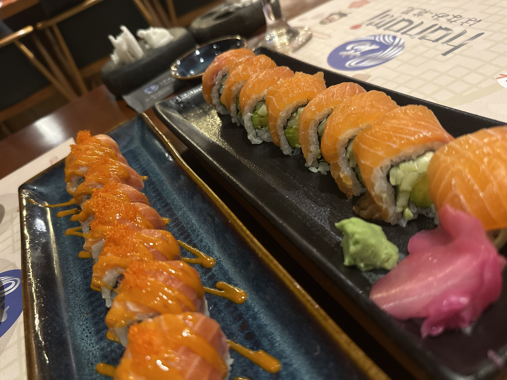 The image presents a close-up, elevated view of two plates laden with sushi rolls, suggesting a meal at a Japanese restaurant. In the foreground, a vibrant blue rectangular plate holds a line of approximately ten sushi rolls. These rolls are topped with a creamy orange sauce and speckled with bright orange tobiko (fish roe), with additional drizzles of the sauce on the plate.
Behind this, on a sleek black rectangular plate, another line of about eight sushi rolls is visible. These rolls feature fresh, thin slices of salmon draped over the top, likely encasing a filling of green avocado and possibly cucumber. Accompanying these rolls on the black plate are a dollop of bright green wasabi and a mound of pink pickled ginger (gari).
The table is set with a patterned paper placemat displaying the stylized English text "Hanamaru" and Japanese characters, indicating the restaurant's name. A reflective glass and a small dark dish are also part of the table setting. The background shows blurred dark wooden furniture, hinting at an indoor restaurant ambiance, likely in the evening given the lighting. The scene captures a prepared meal at a Japanese restaurant located in Nha Trang Ward, Vietnam.