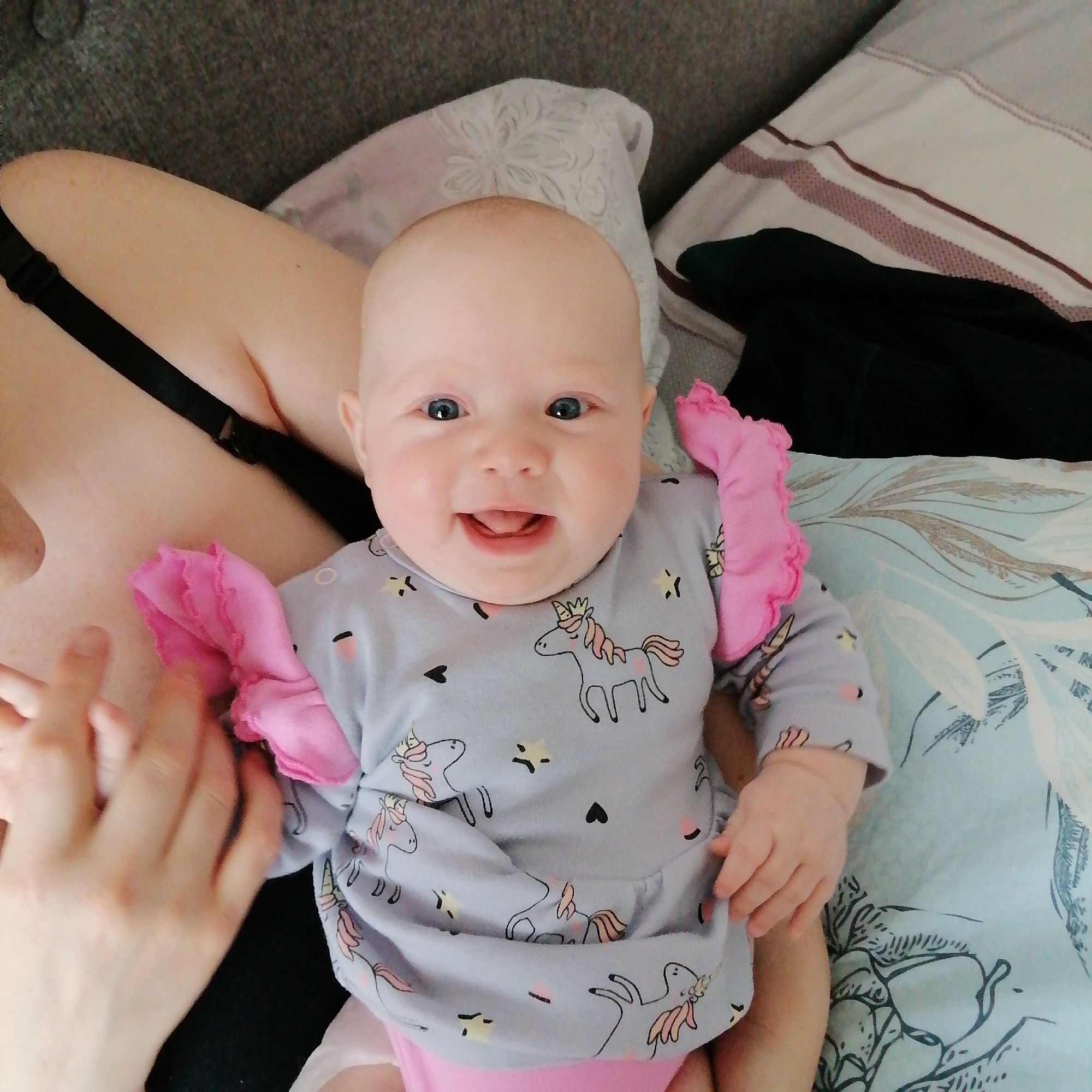 A baby with a unicorn-patterned dress is lying on a surface with a patterned pillow. A hand is holding the baby's hand. The baby is smiling, and its eyes are open. There is also a bra strap visible. The location is Unknown, Unknown.