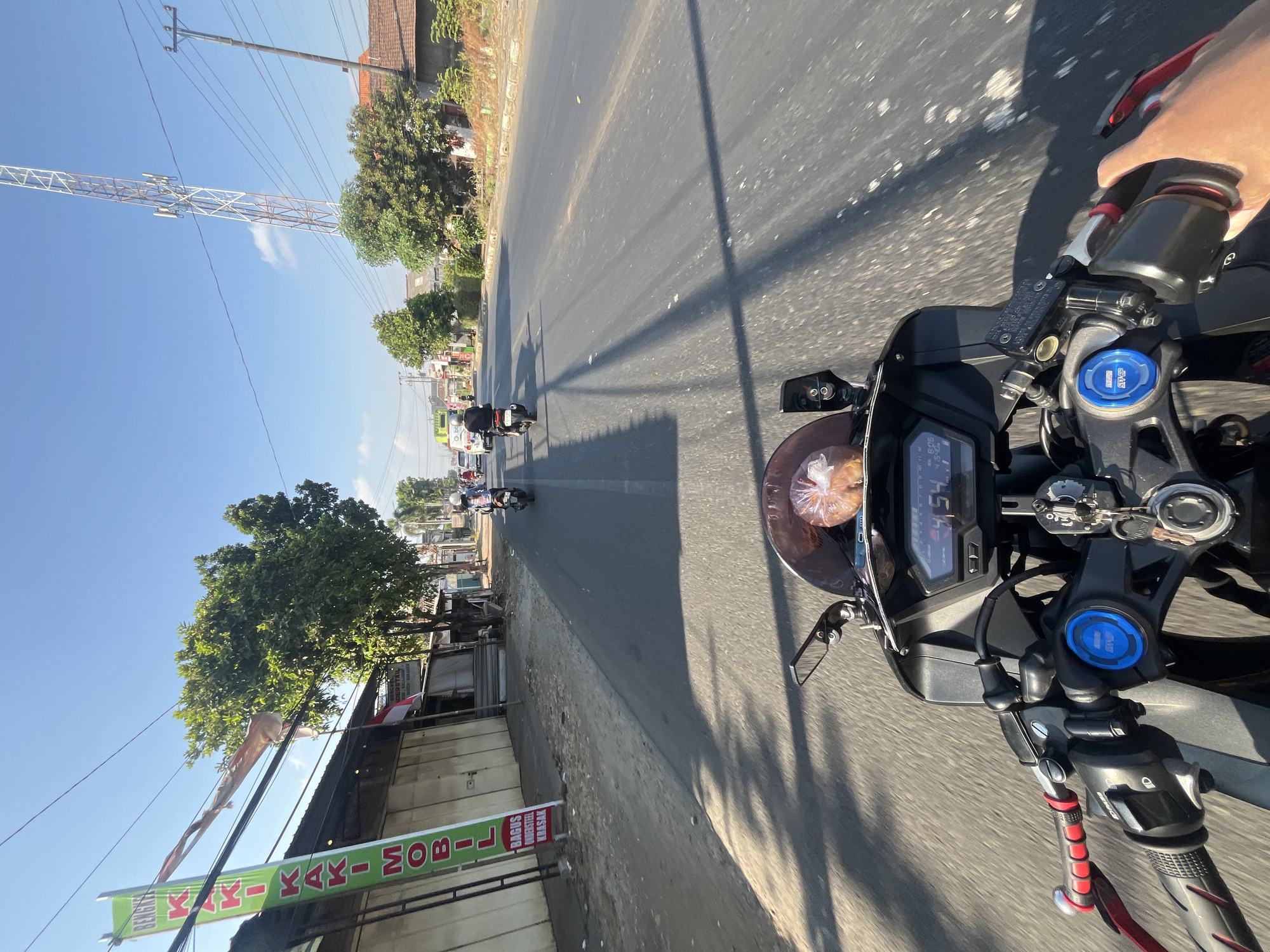The image captures a first-person perspective from a motorcycle traveling on a road in Kalinyamatan, Indonesia. The road is paved and appears to be a two-lane highway with moderate traffic, consisting of other motorcycles and at least one car visible in the distance. The time displayed on the motorcycle's dashboard is 4:54, and the ambient temperature is 30 degrees Celsius.
On the left side of the road, there are several storefronts, one of which has a green banner with vertical text that reads "BENGKEL KAKI KAKI MOBIL" (Car Suspension Workshop). The buildings are low-rise, with a mix of metal roll-up doors and some open structures. Trees line the roadside, and there are power lines and poles present. In the background, a tall telecommunications tower is visible against a clear blue sky.
The motorcycle itself is black, with visible handlebars, controls, and a dashboard displaying speed and time. A rearview mirror is mounted on the handlebars. A plastic bag containing bread or pastries is placed on the front of the motorcycle, just below the windshield. The rider's hands are on the handlebars. The overall environment suggests a sunny day in a semi-urban or peri-urban area of Indonesia.