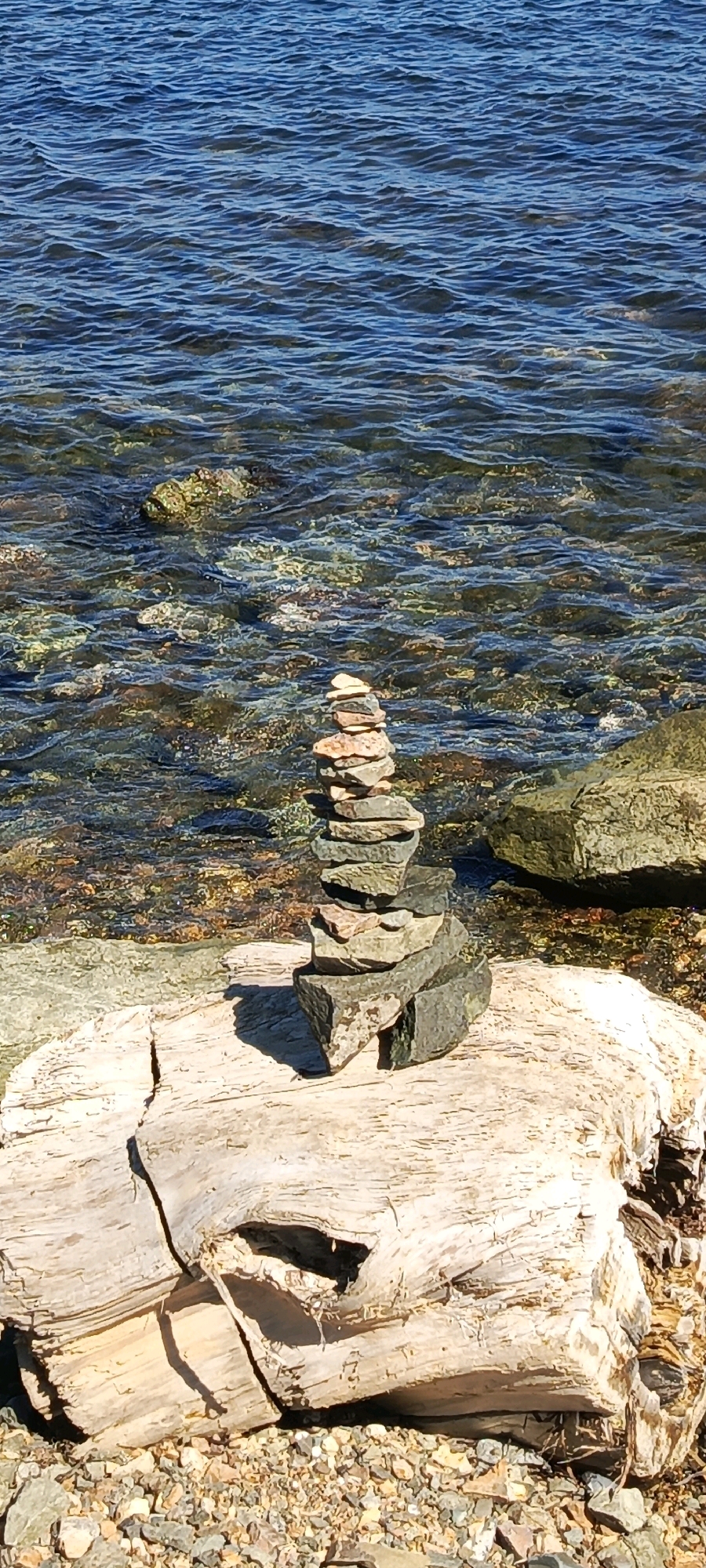 The image shows a cairn, a man-made pile of stones, stacked precariously on a light-colored piece of driftwood. The driftwood rests on a rocky shore, with the clear, blue water of a lake or ocean extending into the background. The water is rippled and shows the colorful stones and sand beneath the surface. The sun casts a shadow from the cairn onto the driftwood.