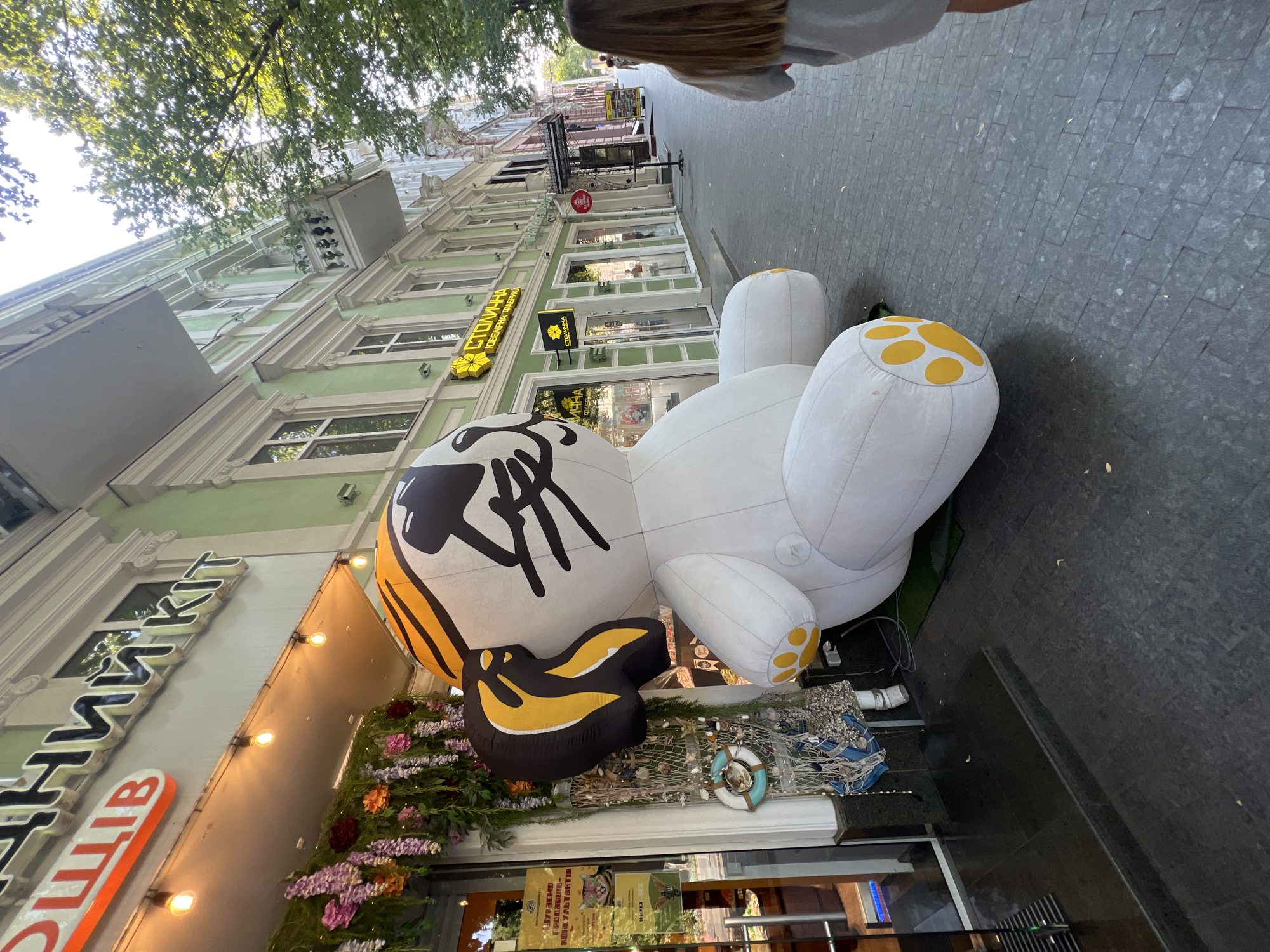 In Odesa, Ukraine, on a sidewalk paved with dark gray square tiles, a large inflatable white cartoon dog stands on its side, facing away from the viewer. The dog has yellow paw prints on its feet and yellow and black markings on its head. It is located in front of a storefront with a green facade, adorned with a sign that reads "Столична Ювелірна Фабрика" (Stolichna Jewelry Factory). To the left of the dog, a glass-fronted shop features hanging floral decorations and a display of nautical items, including a life preserver ring and netting filled with shells. The building behind the shops has a light green exterior with white trim and balconies. Further down the street, other businesses with awnings and signs are visible, and a person with long brown hair is walking away from the camera towards the right side of the image. The overall scene is outdoors, with trees lining the street casting shadows.