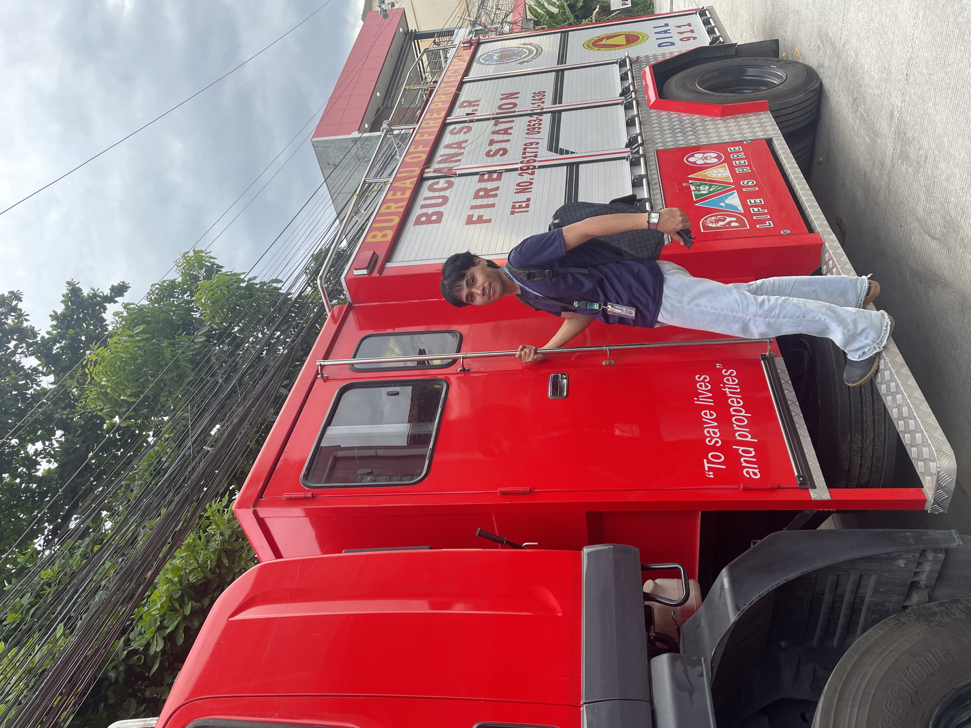 A young Filipino man with dark hair and light skin stands beside a bright red fire truck. He is wearing a dark blue t-shirt, light blue jeans, and a black backpack. The fire truck has "BUREAU OF FIRE PROTECTION BUCAANA S.I.R. FIRE STATION" written in large letters on its side, along with a phone number and a logo. Below that, it says "To save lives and properties." A colorful "DAVAO LIFE IS HERE" logo is also visible on the truck. The man is smiling as he looks at the camera. The background shows trees, overhead electrical wires, and a cloudy sky. The scene appears to be in a street setting in Davao City, Philippines, given the "DAVAO LIFE IS HERE" branding on the fire truck.