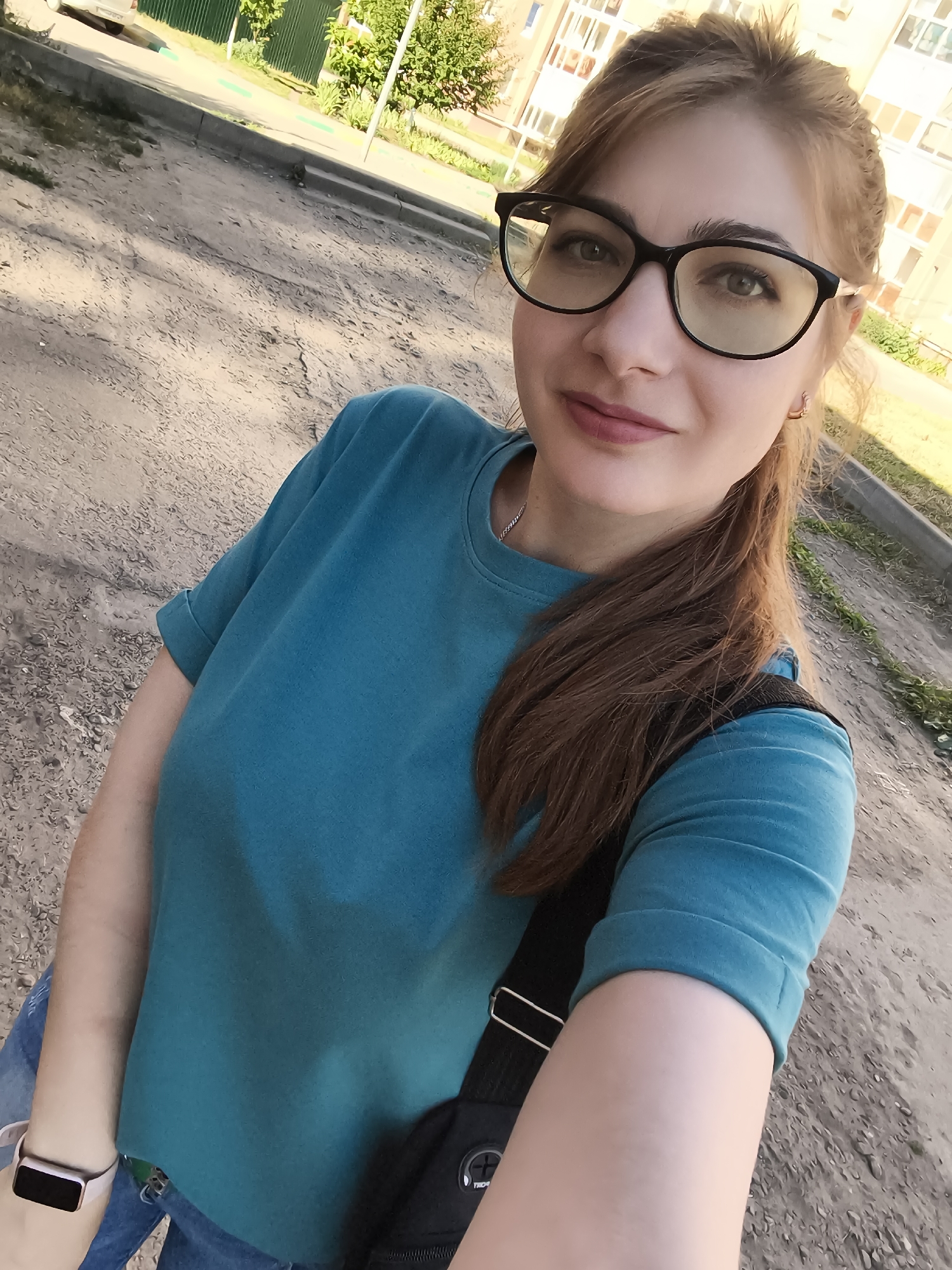 A woman with light brown hair pulled back and wearing black-rimmed glasses and a teal t-shirt takes a selfie outdoors. She is wearing blue jeans and a smartwatch on her left wrist. A black bag is slung across her right shoulder. The background shows a dirt path, a sidewalk, green grass, trees, and a building with multiple windows. The scene appears to be in an urban environment.