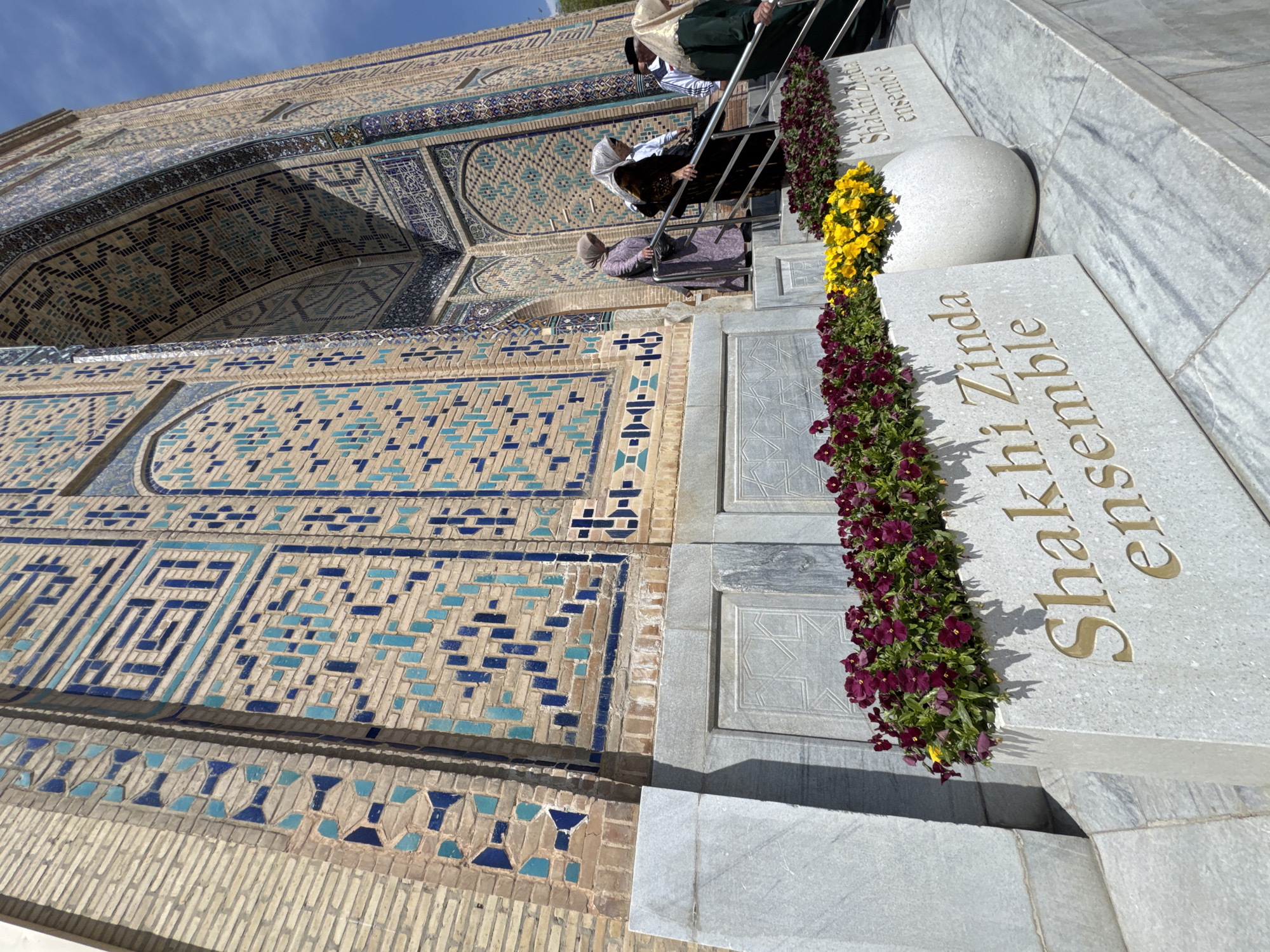 The image shows a section of the Shah-i-Zinda ensemble in Samarkand City, Uzbekistan. The photograph is taken from a low angle, emphasizing the architectural details and decorative elements.
On the left, there is a wall covered in intricate tilework featuring geometric patterns in blue and brown. The tiles appear to be arranged in various designs, creating a visually complex texture. The wall seems to be part of a larger structure, possibly a tomb or mausoleum, given the location.
On the right, there is a stone staircase leading upwards. The steps are made of white marble or a similar light-colored stone. The steps are adorned with planted flowers in red and yellow.
People are partially visible on the staircase, seemingly descending. They are dressed in various colors and patterns and can only be seen from the upper torso up.
The words "Shakhi Zinda ensemble" are written on the side of the stone steps in gold letters.