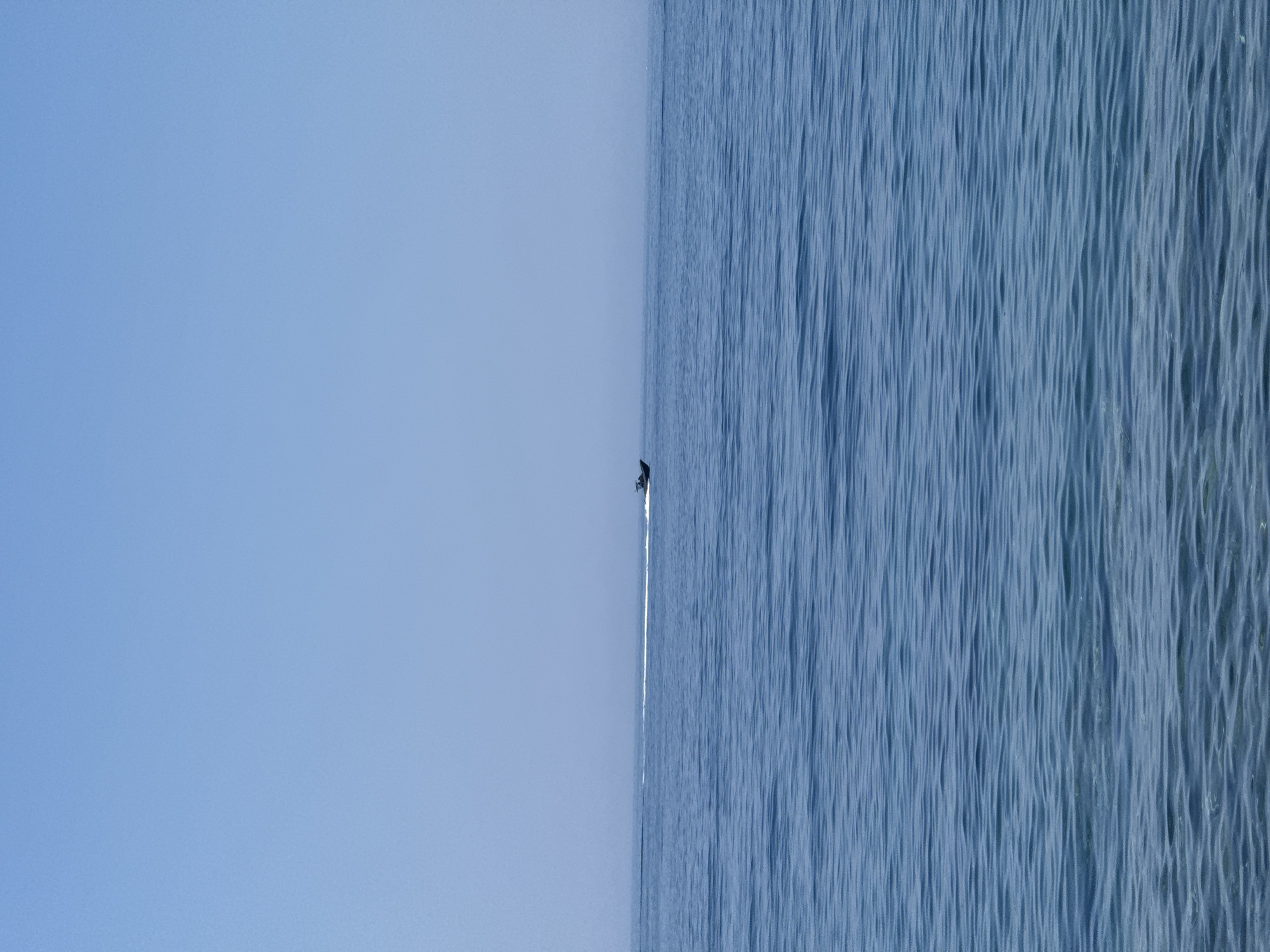A wide shot captures a vast expanse of blue ocean under a clear, pale blue sky. In the mid-ground, a small, dark boat is moving from left to right, leaving a white wake behind it. The water's surface is textured with gentle ripples, creating patterns of light and shadow. The horizon line is faintly visible, separating the sea from the sky, with a slight atmospheric haze obscuring distant details. The scene is devoid of any landmass or structures, suggesting a view from offshore. The location is identified as the waters near Sveti Stefan, Montenegro.
