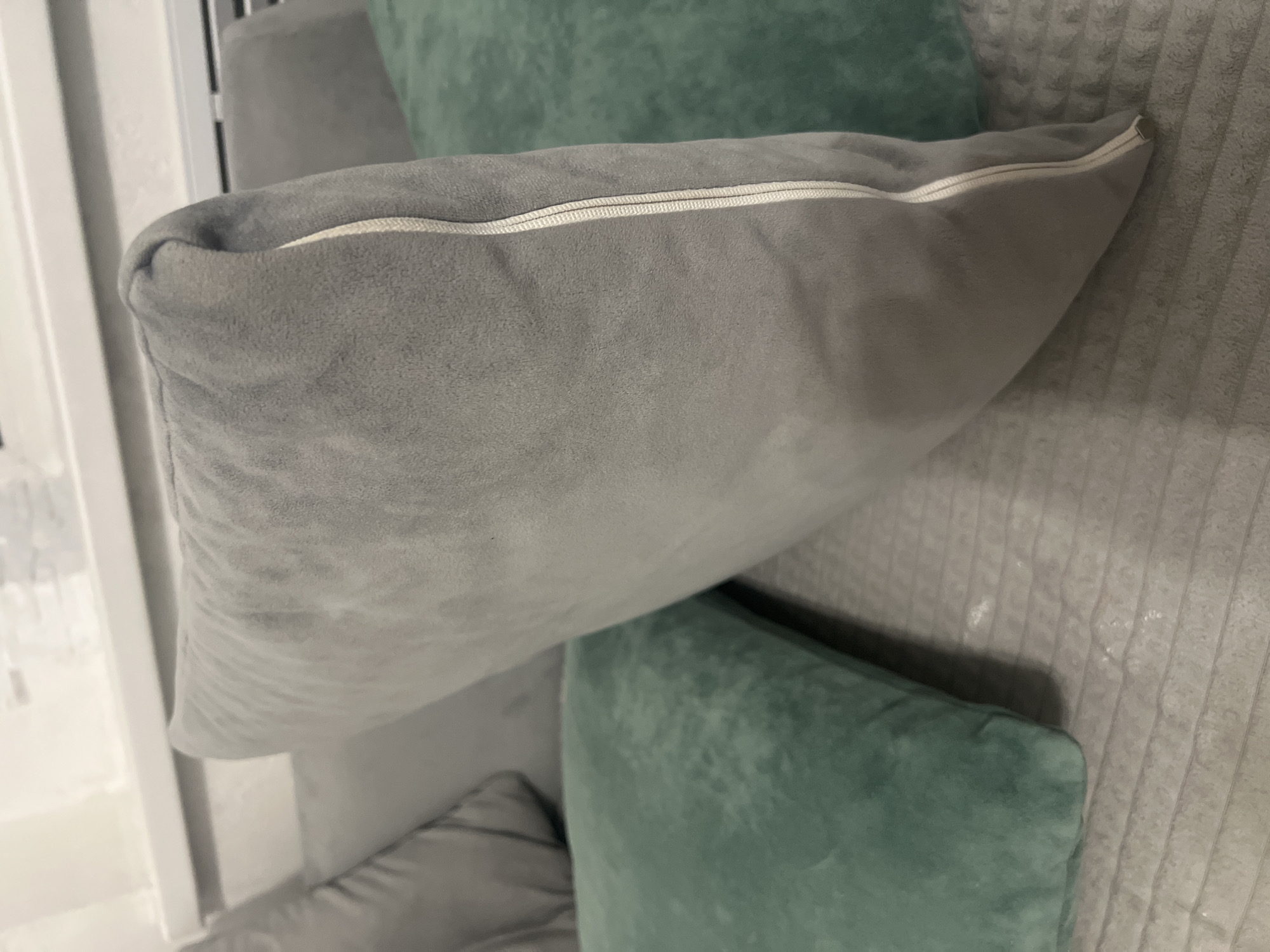 The image shows a close-up of several pillows on a couch. In the foreground, a large gray pillow with a visible zipper runs along its side is angled towards the right. To the left of the gray pillow, a teal-colored pillow is visible. Behind these, another gray pillow is partially obscured. The couch itself appears to be upholstered in a textured gray fabric. In the background, a white ledge with a horizontal slat design is visible, suggesting this may be a windowsill or a similar architectural feature. The lighting in the image is soft, creating a slightly diffused atmosphere. No people are present in the scene. The location of Veteran-2, Russia is not depicted or suggested by any elements in the image.