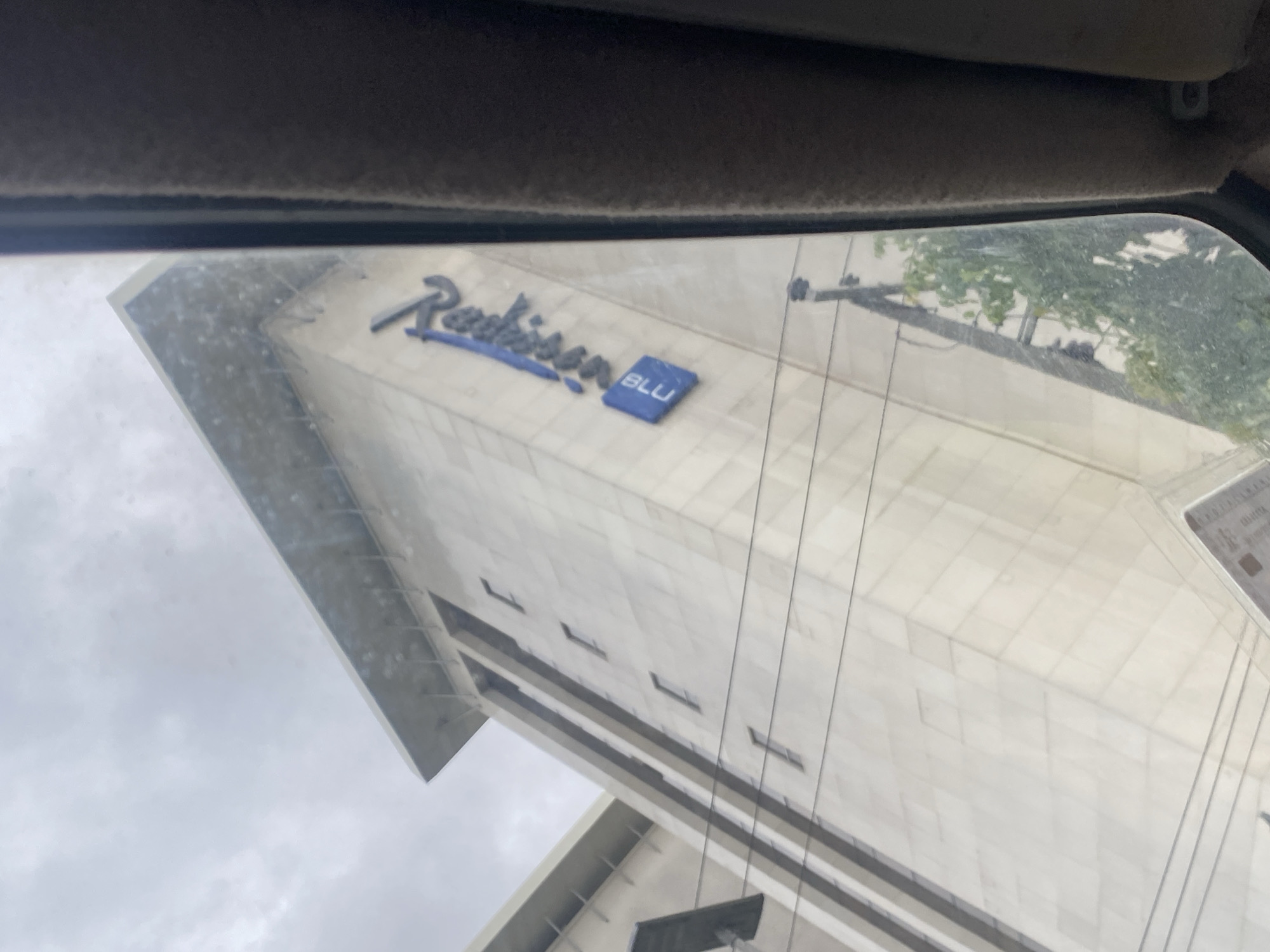 This is a low-angle, slightly blurry exterior shot of the Radisson Blu hotel in Ikeja, Nigeria, taken from inside a vehicle, likely through the windshield and side window. The building's facade is light-colored concrete, with a geometric, angular design. Several narrow, vertical windows are visible. A prominent blue Radisson Blu logo is mounted on the building's surface, with the word "Radisson" in stylized script and "BLU" in a blue square below it. Electrical wires crisscross the foreground, obscuring parts of the lower section of the building and some greenery at its base. The sky above is overcast and grey. The interior of a car, including the side pillar and fuzzy headliner, is visible along the right edge of the frame, indicating the viewpoint.