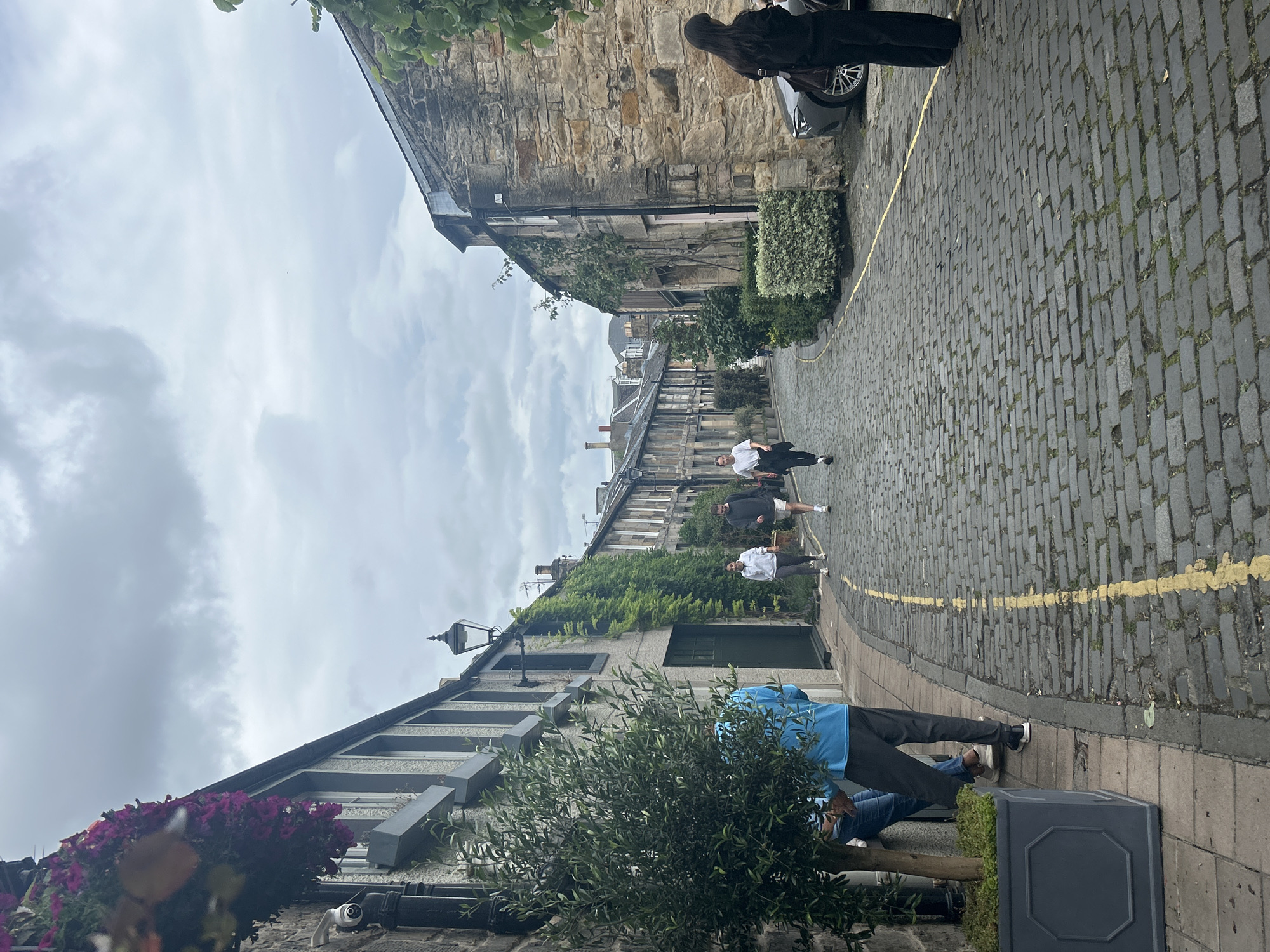 The media file depicts a narrow, cobblestone street in Edinburgh, Scotland, under an overcast sky. Several people are walking or standing on the street and sidewalk.
In the foreground, a person in a bright blue hooded jacket is seated, facing away from the camera, with another person in jeans beside them. Further up the street, a group of three people are walking away from the camera. One person is wearing a white shirt and shorts, another is in a black top and shorts, and the third is in a dark top and shorts. Near the top of the image, a person with long dark hair, wearing black pants and a black top, is standing next to a car.
The buildings lining the street are constructed from stone, with some featuring ivy growth on their facades. One building has a prominent lamppost. The street is marked with a yellow line along the curb. The weather appears to be cloudy, suggesting it might be a cooler day. The overall scene conveys a sense of everyday activity in an urban environment. No visible text is present in the image.