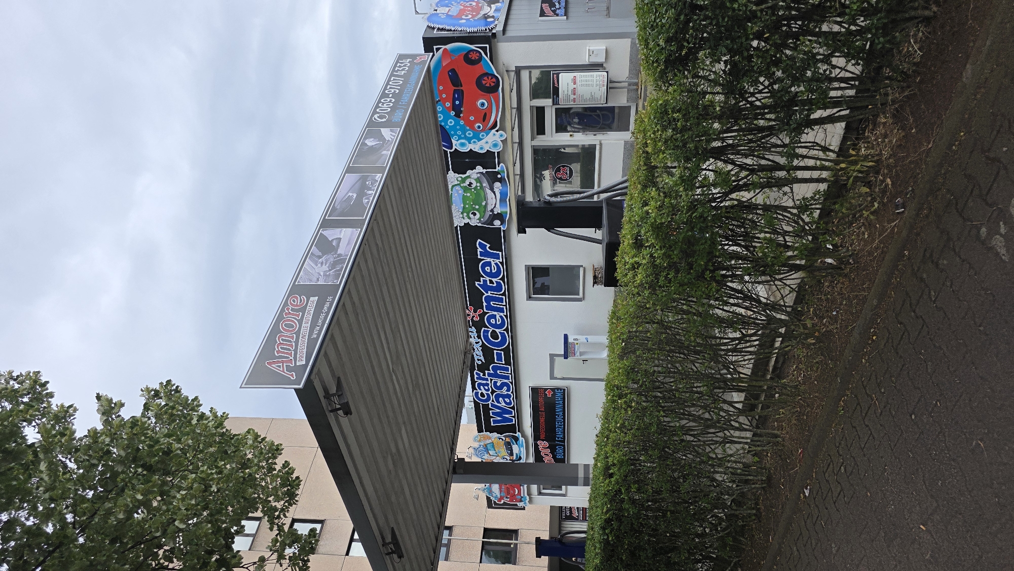 This is an eye-level, full shot of a car wash center called "Amore Car Wash-Center" on a cloudy day. The establishment is housed in a white building with a gray roof overhang. A well-maintained green hedge runs along the front of the building, separating it from a paved walkway. The car wash features cartoonish graphics of a green and red car with faces. A sign above the overhang reads "Amore PROFESSIONELLE AUTOIPFLEGE" with a website address and a phone number. The building also has windows and doors, and to the right, a blue pole with a vacuum cleaner is visible. In the background, a multi-story building and a tree with green foliage can be seen.