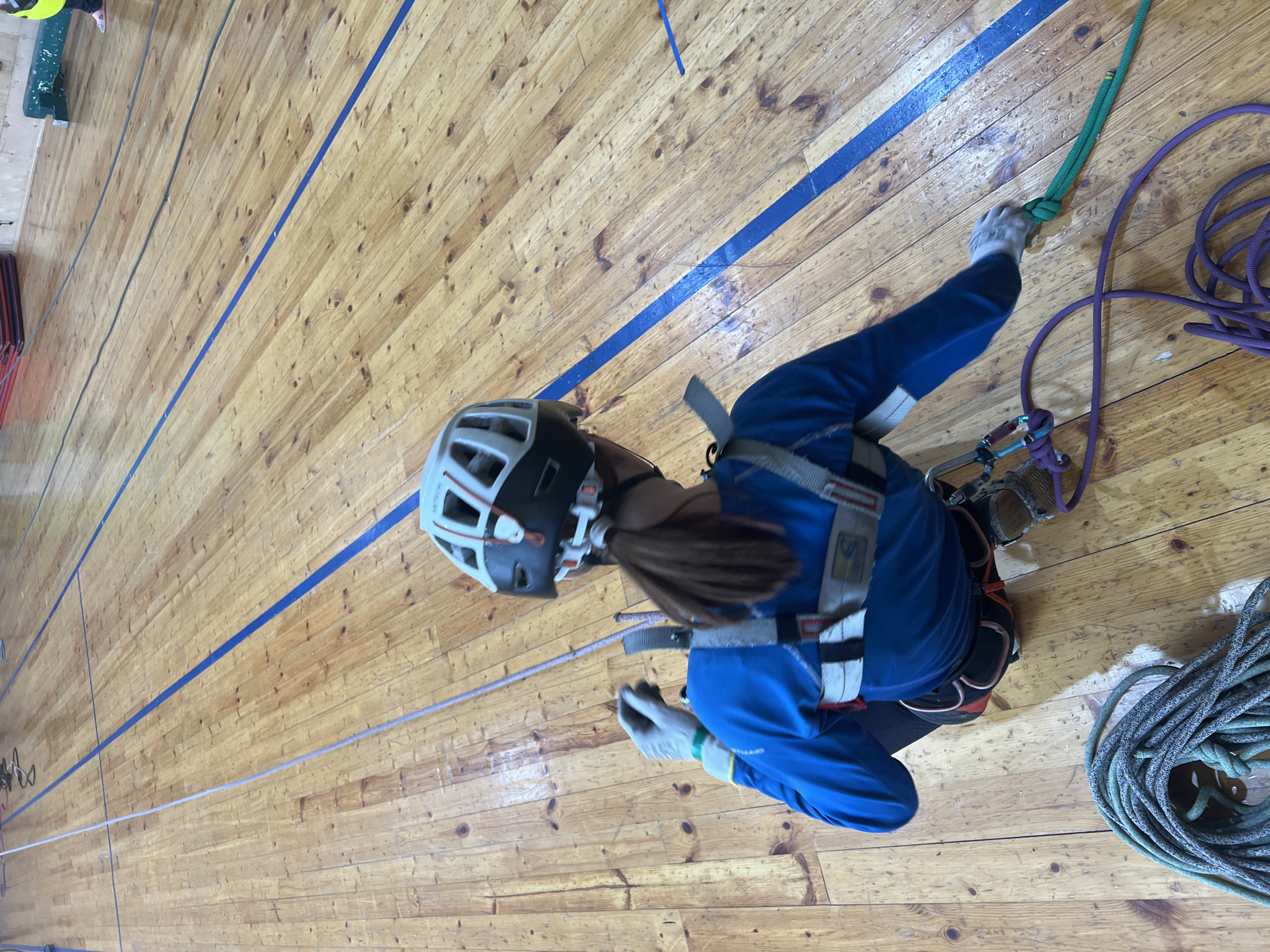 A woman is indoor rock climbing in Chaikovsky, Russia. She wears a climbing helmet, harness, gloves, and a blue jacket. She holds onto ropes and is in the process of climbing a tall wooden wall with blue stripes. The wall appears to be part of an indoor climbing facility, as coils of ropes lay on the floor next to the wall.