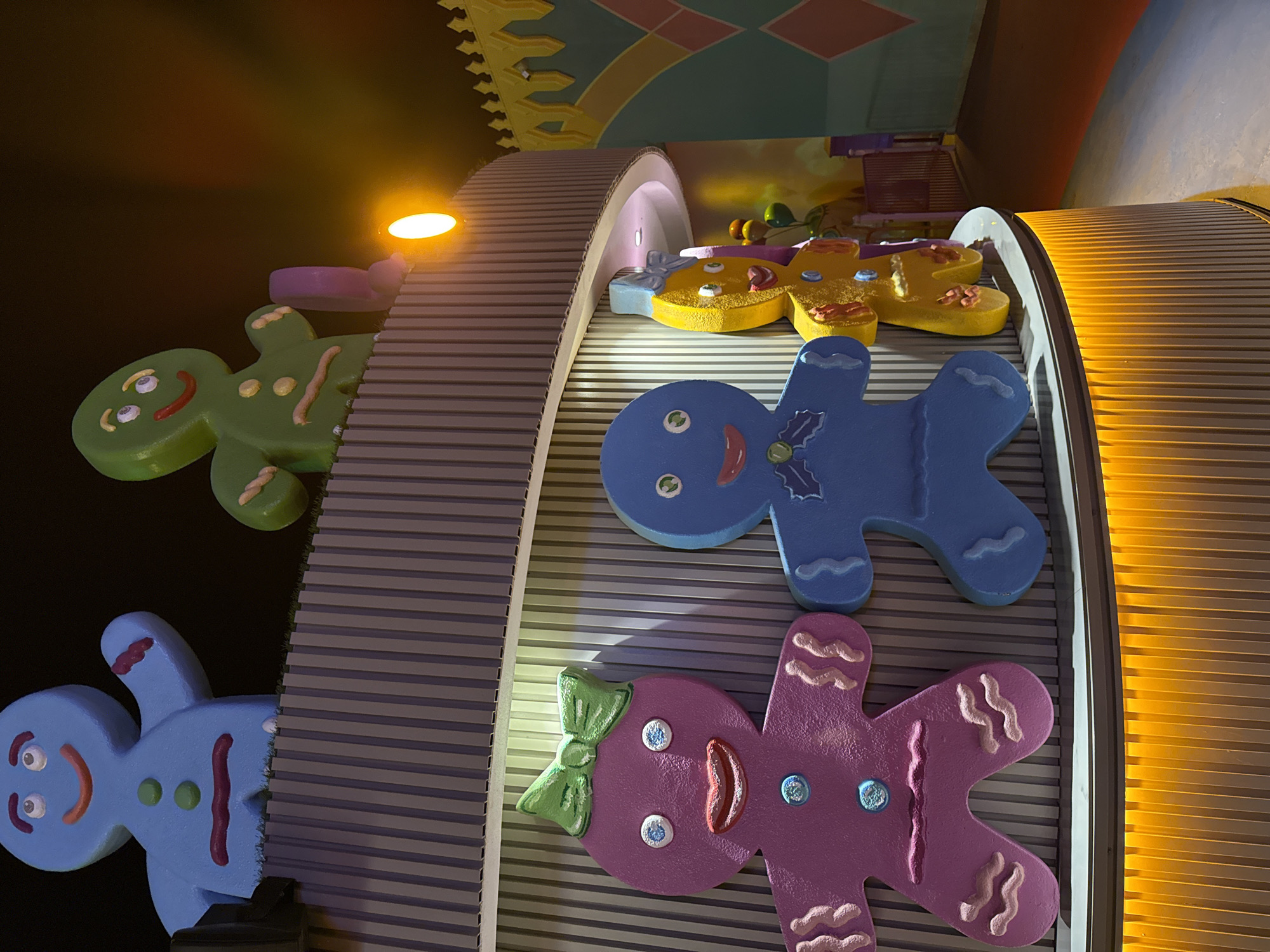 This image captures a whimsical display of gingerbread figures in a dimly lit indoor environment, possibly an amusement park or entertainment venue in Bodrum, Turkey. In the foreground, three large gingerbread figures are arranged side-by-side: a pink female figure with a green bow on her head, a blue male figure, and a yellow figure. Behind them, partially visible, are two larger gingerbread figures, one light blue and the other green, positioned on a higher level. The figures are stylized with large, circular eyes, smiling mouths, and button-like decorations. The pink figure has a green bow and blue buttons, while the blue figure has a blue ribbon design and a green button. The yellow figure has red decorations on its arms and chest, and a small blue bow. The gingerbread figures are set against a backdrop of vertical ribbed walls. The lower part of the structure features illuminated yellow ribbed panels. In the background, a glimpse of a colorful, geometric mural and a purple railing is visible, suggesting a themed area. The overall atmosphere is playful and festive, with warm lighting illuminating the scene.