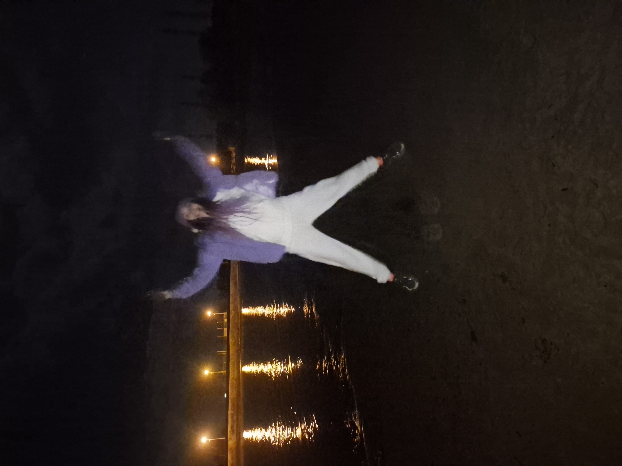 At night, an individual is lying on their back on dark ground near a body of water in Noviy, Russia. The person, wearing a white outfit and a purple jacket, is positioned with arms extended and legs spread, mimicking the shape of a snow angel. The dark ground is textured. In the background, lights from a structure over the water reflect onto the surface, indicating the presence of a pier or similar construction. The overall environment appears dark with limited visibility. The surrounding area is mostly darkness.