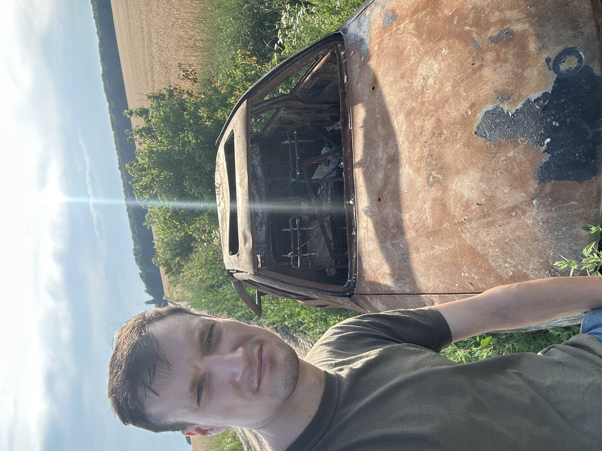 A man with short brown hair and a light complexion stands in the foreground, taking a selfie. He is wearing a dark olive-green t-shirt and blue jeans. To his right is a heavily damaged, burned-out car. The car's roof is open, and the interior appears gutted and charred. The exterior of the car is rusted and discolored, with significant areas of peeling paint and exposed metal.
The scene is set in a rural area. Behind the man and the car, a dirt road is visible, flanked by green grass and vegetation. Further in the background, a field of ripe wheat or barley stretches towards a dense line of trees, forming a dark green border against a pale blue sky with scattered clouds. A faint lens flare emanates from the upper left quadrant of the image. The overall impression is one of destruction and desolation in a natural landscape. The location is identified as Makiv, Ukraine.