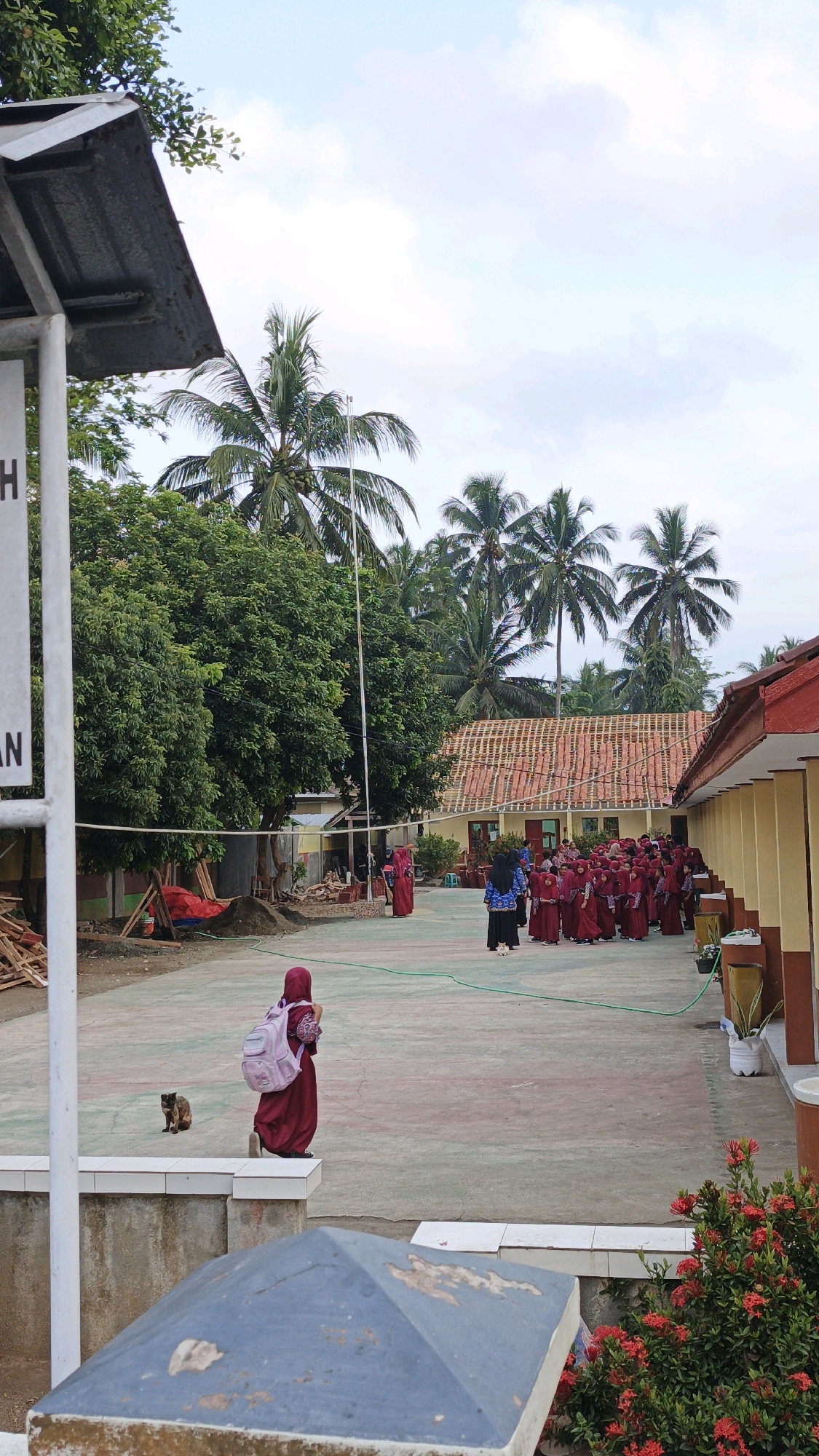 The image depicts a courtyard likely belonging to a school in Cibenda, Indonesia, under a cloudy sky with some palm trees and lush green trees visible in the background. In the foreground, a young student wearing a maroon hijab and a pink backpack walks away from the camera, facing a group of other students gathered in the courtyard. The students, predominantly girls, are dressed in maroon uniforms and appear to be lining up or congregating. One adult, also in a dark headscarf and maroon top, stands facing the students. To the left, a small, dark cat is seated on the ground, observing the scene. The courtyard surface is paved with a reddish and greenish patterned material. On the right side of the image, a yellow building with red trim runs along the courtyard, with a few potted plants near its entrance. Construction materials, including lumber and a red tarp, are visible in the background to the left, suggesting ongoing work. The overall impression is of a school day in progress, possibly during an assembly or break time.