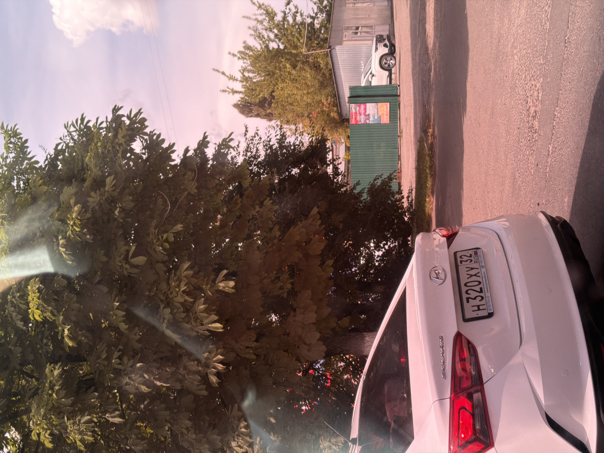 The image shows a white Hyundai Solaris sedan parked on an asphalt surface. The car's rear is visible, including its taillights and license plate, which reads "H 320 XY 32". The car is positioned in what appears to be a parking lot or side street in Bryansk, Russia, indicated by the license plate's regional code. In the background, there is a large tree with dense foliage, a green corrugated metal fence, and a building with a corrugated metal roof. Another white SUV is parked behind the fence. The sky is visible with some clouds and a pinkish hue, suggesting either sunrise or sunset. The overall scene is ordinary, depicting a parked car in an urban or suburban setting.