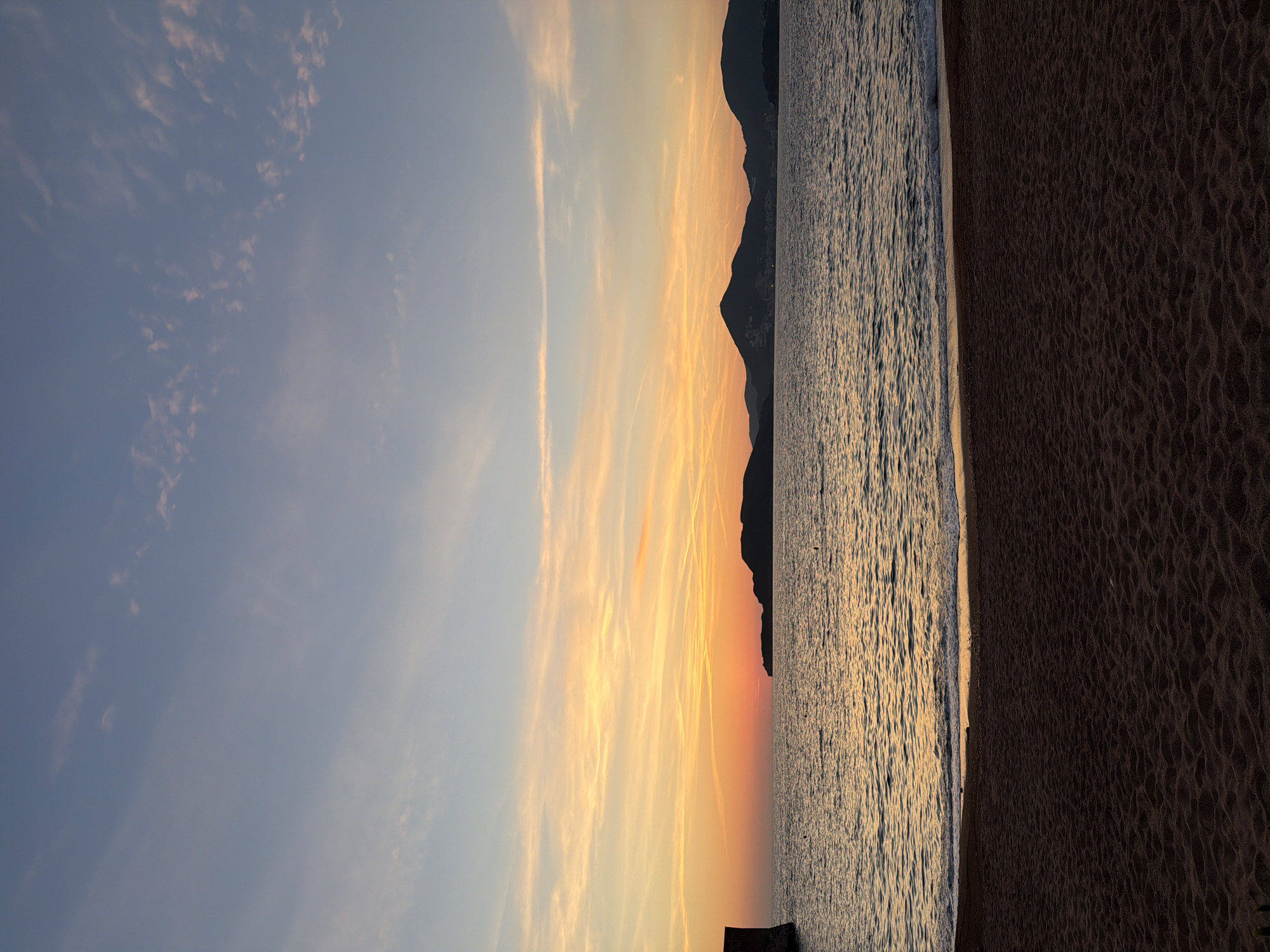 The image depicts a sunset over a body of water and coastline, presumably captured from a beach in Sveti Stefan, Montenegro. The sky, occupying approximately the upper two-thirds of the frame, transitions from a light blue at the top to vibrant orange and yellow hues near the horizon, indicative of a sunset. Streaks of wispy clouds are visible, particularly in the warmer-toned areas.
Below the sky, the sea extends across the middle of the frame, reflecting the sunset colors in a shimmering, golden-orange glow that diminishes towards the foreground. Small waves are visible, breaking gently at the shoreline. Several small, dark buoys are sparsely scattered across the water's surface in the mid-ground.
Along the horizon, a silhouette of multiple dark mountains and hills stretches from the center to the right. A prominent, darker landmass is visible closer to the center-right, obscuring a portion of the setting sun. On the far left, a dark, angular structure, possibly a part of a wall or building, is partially visible, extending from the left edge into the water.
The foreground consists of a dark sand beach, appearing damp near the waterline and displaying a textured, uneven surface with small indentations. The demarcation between the water and the sand is clearly visible, with a thin line of white foam from the breaking waves. There are no identifiable people or animals in the scene.