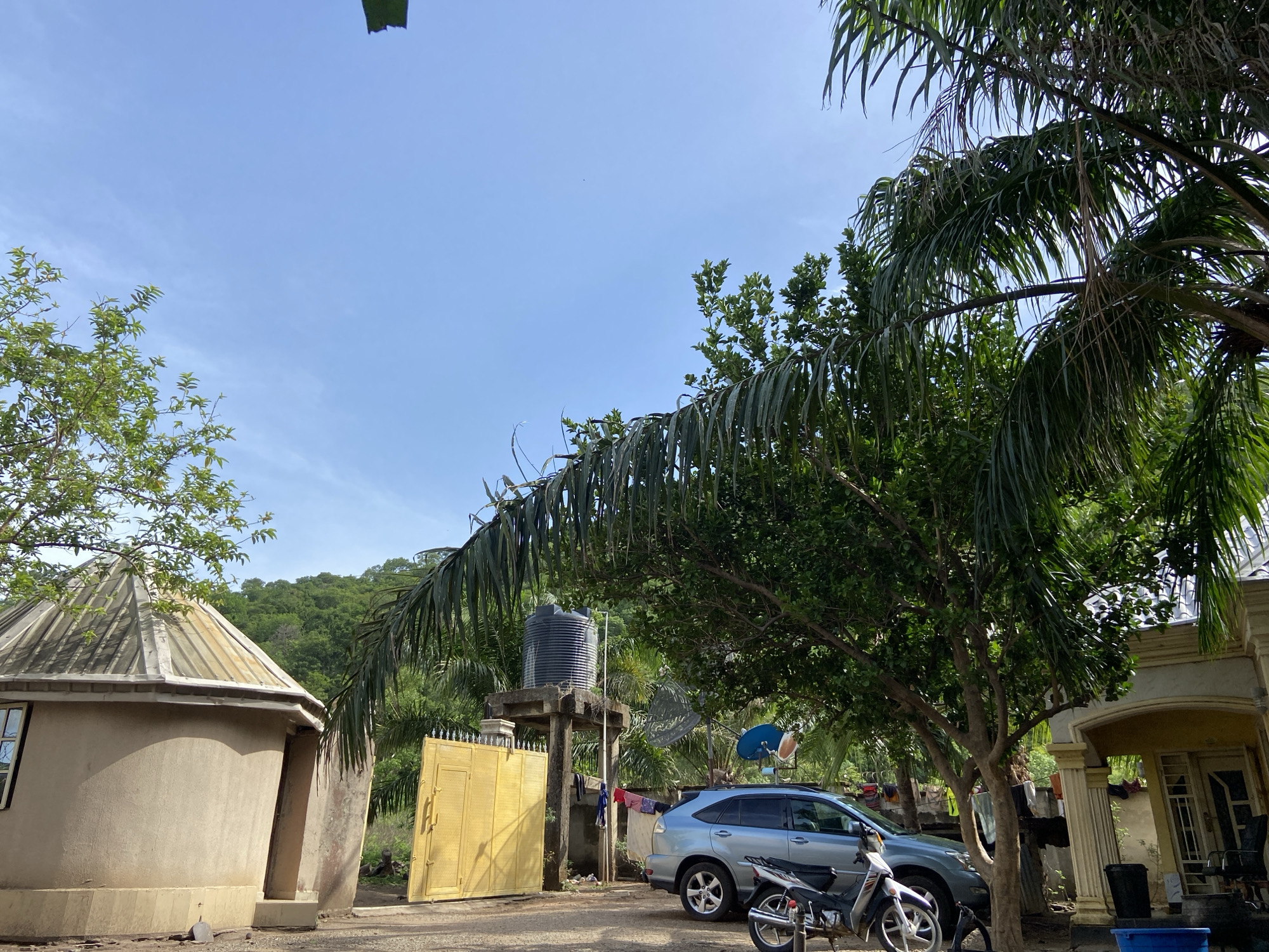 The image shows a residential area in Mararaba, Nigeria. A small, round building with a conical roof is on the left. A yellow metal gate stands in front of a water tank supported by a concrete structure. A light blue SUV and a motorcycle are parked nearby. A palm tree and other trees provide shade over the area. Clothes are hung on a line near the gate. A portion of a larger house is visible on the right. A forested hill forms the backdrop of the scene. The sky is mostly clear with light blue.