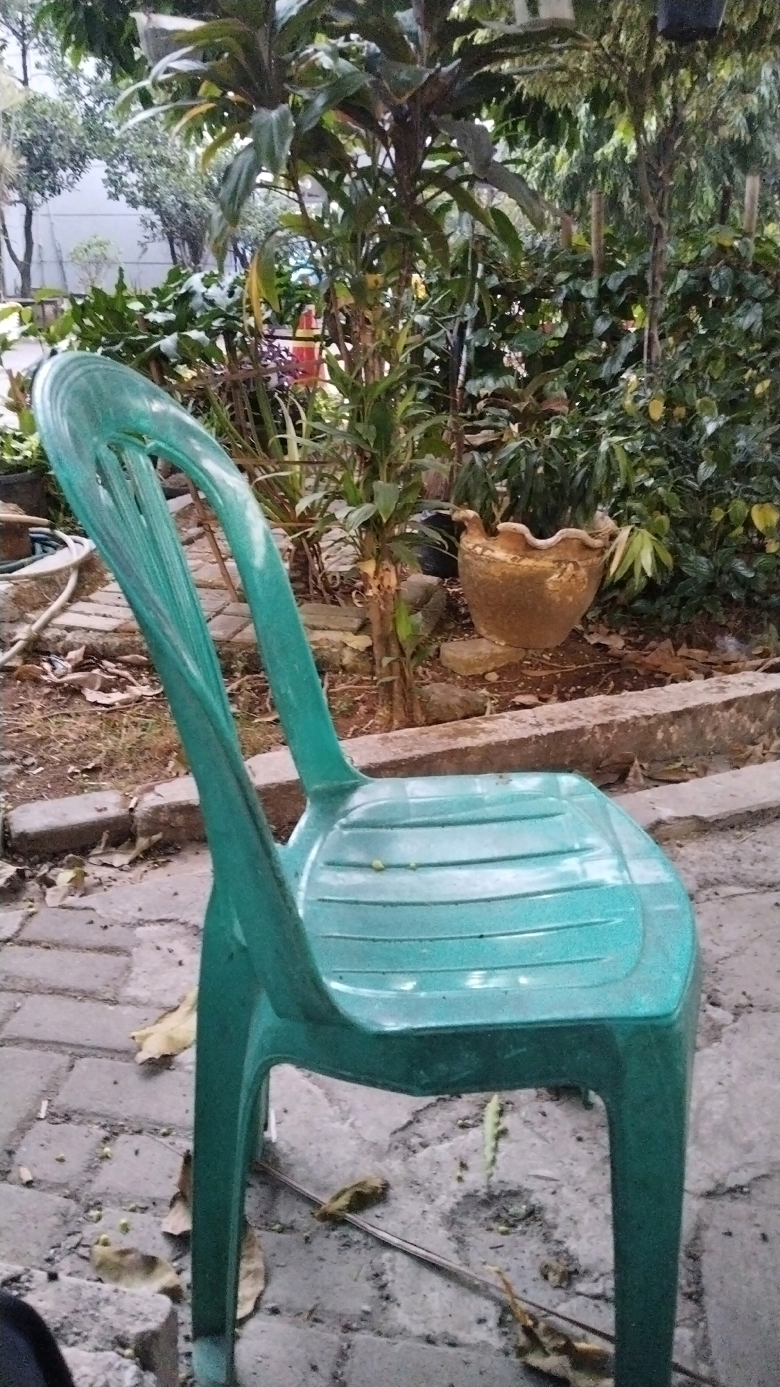 The image shows a green plastic chair in an outdoor setting. The chair is in the foreground, positioned at an angle. Behind the chair, there is a garden with various plants, trees, and a decorative pot. The ground is paved with bricks and scattered with leaves and debris. The overall scene appears to be a residential or communal outdoor area. The context of Bekasi, Indonesia is not visually evident from the image itself.