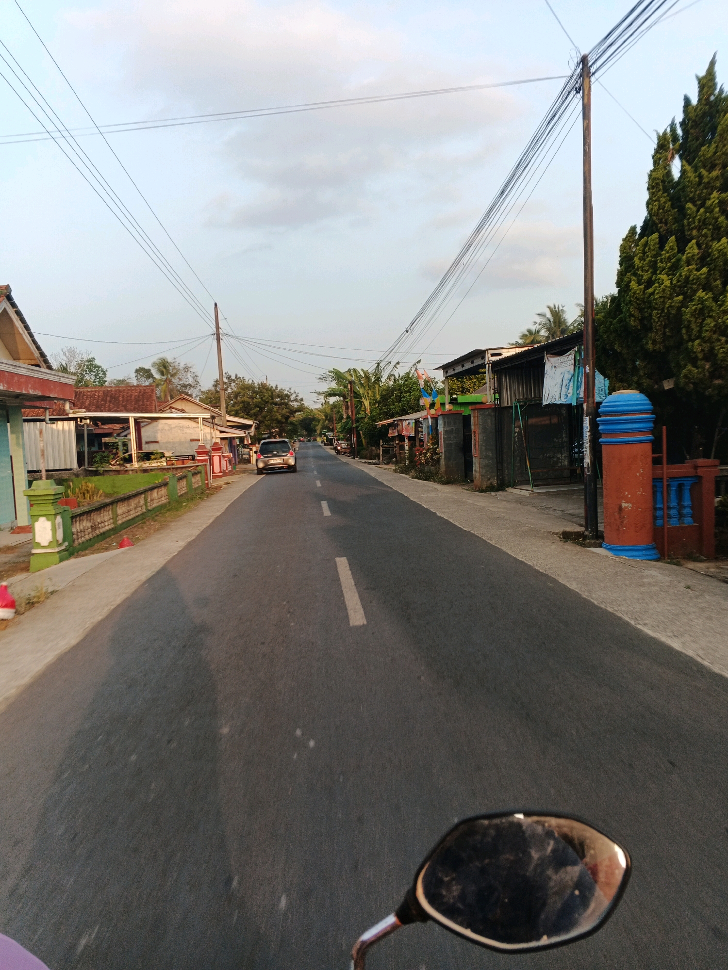 The image captures a straight asphalt road in the city of Pucung Kidul, Indonesia. The road is flanked by houses and fences on both sides, with power lines and utility poles overhead. A silver car is visible driving away from the camera, and the reflection of the photographer's motorbike's mirror is in the foreground. The sky is a pale blue with scattered clouds, suggesting it is either early morning or late afternoon. The overall scene depicts a typical suburban street in Indonesia.