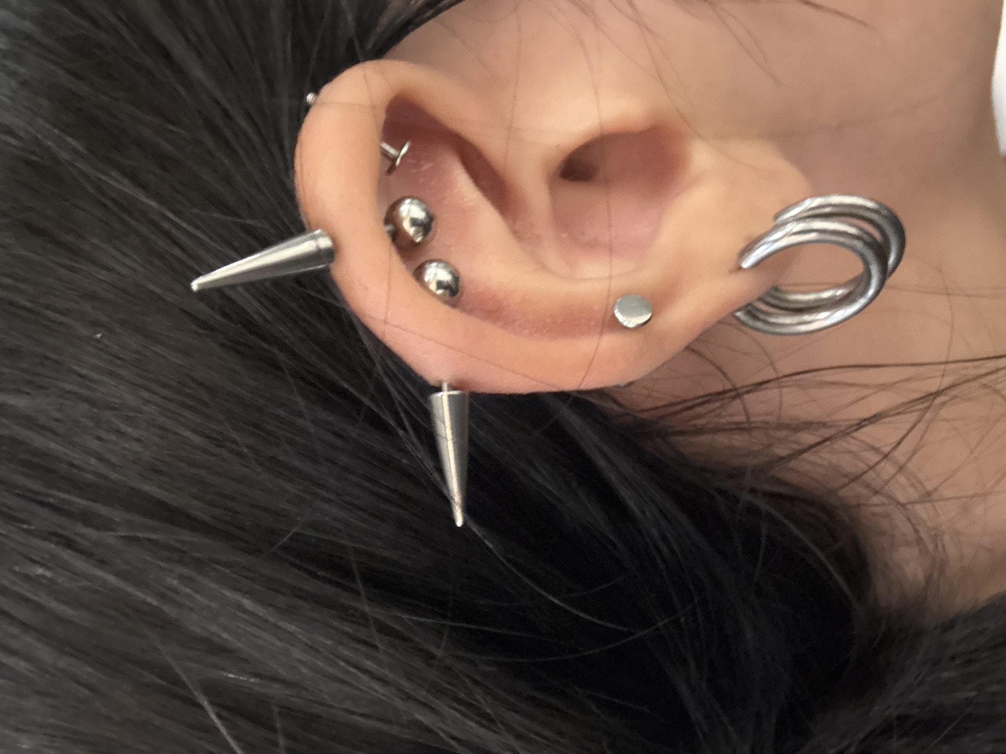 A close-up, eye-level shot shows a person's left ear with multiple piercings and jewelry. The hair is dark and appears to be styled away from the ear. Visible piercings include two conical spike earrings on the upper helix, three small ball stud earrings along the inner ear cartilage, a flat disc stud on the antihelix, and a triple hoop earring in the lower earlobe. The background is out of focus, suggesting an indoor setting.