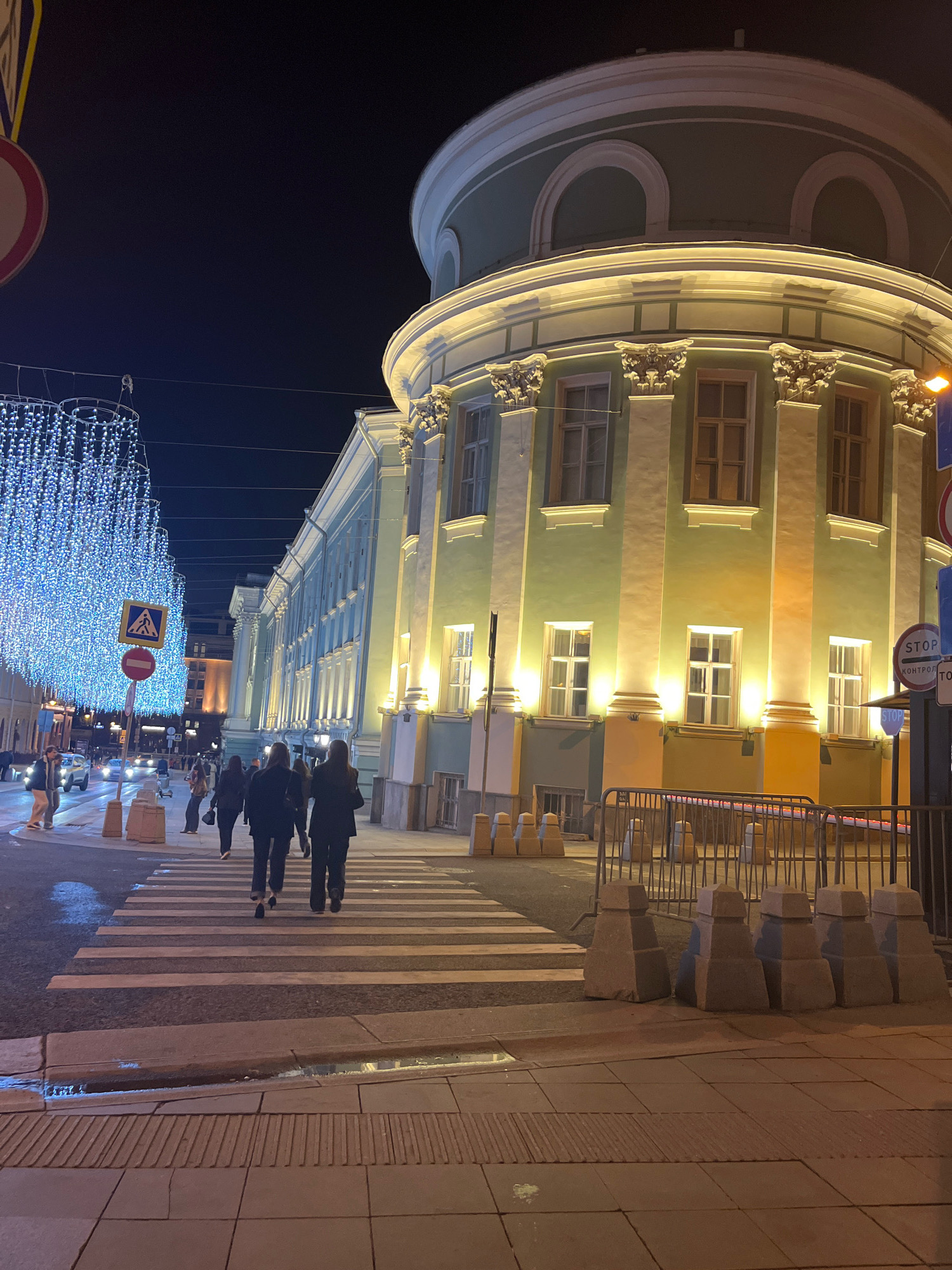 The image shows a nighttime view of a street in Moscow, Russia, possibly in the Arbat District, decorated with bright blue string lights. A distinctive building with a curved facade and classical architectural elements, illuminated by warm yellow lights, dominates the right side of the frame. People are crossing a pedestrian walkway in the foreground, with several cars visible in the background. Barriers and concrete bollards are placed along the sidewalk, suggesting a possible event or security measure. The overall scene is urban and festive, with the lights creating a prominent atmosphere.