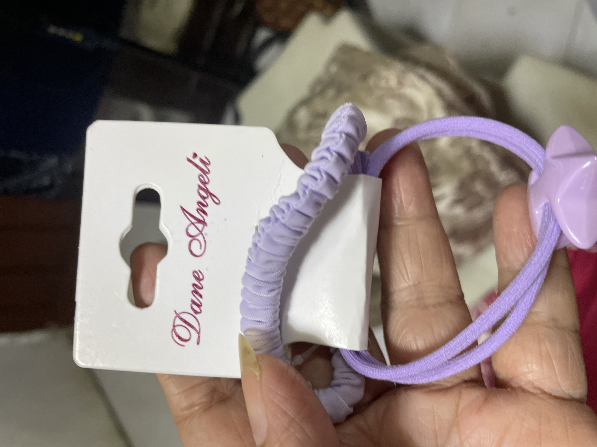 A close-up image shows a hand holding a white cardboard hang tag with two light purple hair accessories. One hair tie is a scrunchie-style elastic with gathered fabric, while the other is a simple elastic band adorned with a flat, star-shaped plastic embellishment, both in the same light purple shade. The hand, which exhibits visible wrinkles and a slightly discolored fingernail, holds the items against a softly blurred background that suggests an indoor setting with various indistinct textures, possibly fabrics. The activity depicted is the presentation or display of these hair accessories. Visible text on the white hang tag, printed in a stylized, dark red or purple cursive font, reads "Dane Angeli." The tag also features a standard punch-out slot, typically used for hanging items on a display rack. The setting is indoors, with no specific time of day or weather discernible from the image itself, but the provided context places the location in Davao City, Philippines.