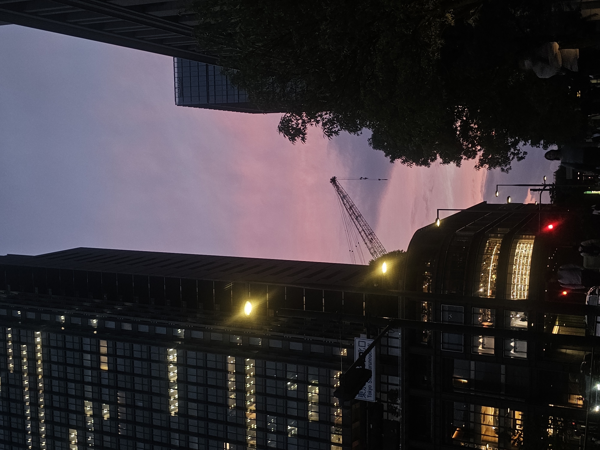 The image depicts a twilight scene in Chiyoda, Japan, with the sky displaying a gradient of purple and pink hues. Dominating the left side of the frame is a tall, dark skyscraper with numerous illuminated windows, suggesting office spaces or residences. To the right, another building is partially visible, its facade silhouetted against the sky. In the mid-ground, a construction crane extends upwards, its metallic structure stark against the colorful sky. Trees with dark foliage are present on the right side of the image, obscuring some of the cityscape. In the foreground, streetlights cast a warm glow, illuminating a street with pedestrian and vehicle traffic. Several individuals are visible, some walking on the sidewalk, others in vehicles. A traffic light shows a red signal. A sign with Japanese text, "Tokyo Sta. Marunouchi Exit," is visible on a pole. The overall atmosphere is that of a bustling urban environment transitioning from day to night.