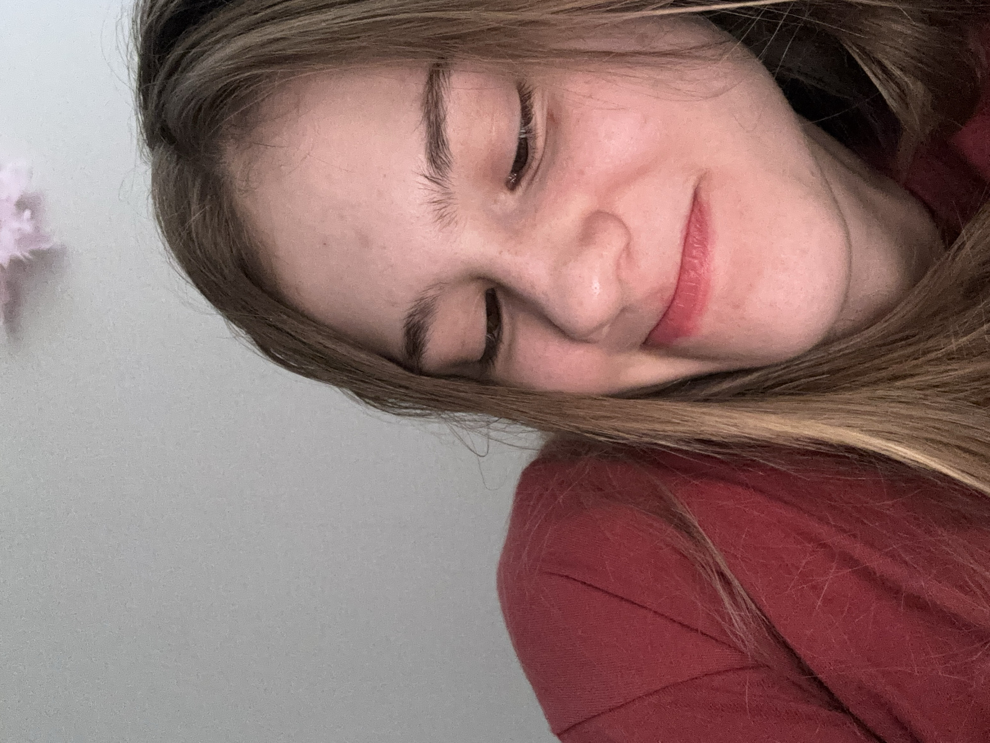 The image is a close-up, indoor portrait of a young person with blonde hair, wearing a red top. Their eyes are closed, and they have a slight smile. The background is a plain, light-colored wall. The overall lighting suggests a casual, informal setting. There is no visible indication of the city of Vsevolozhsk, Russia, or any specific urban environment.