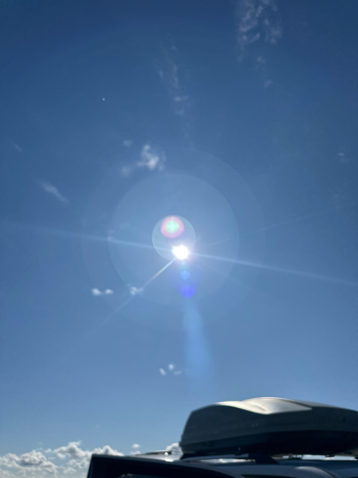 The image shows a bright sun with lens flare in a clear blue sky. There are a few wispy clouds scattered across the sky. In the foreground, the top of a dark-colored cargo box on the roof of a vehicle is visible. The image does not provide any specific information or visual cues that would allow for the identification of the location in the city of Tsabelevka, Kazakhstan.