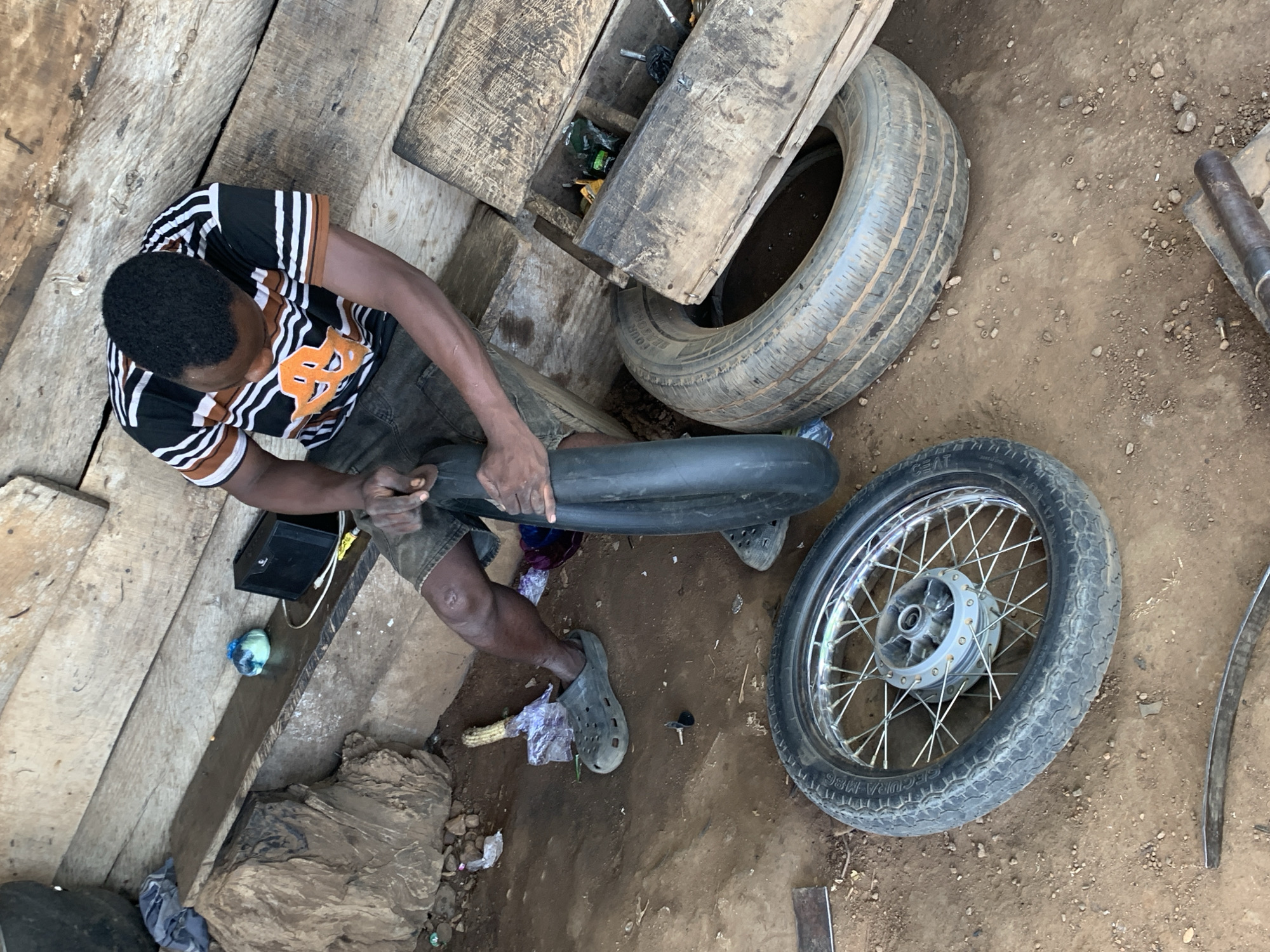 The image captures a top-down view of a person in Kabba, Nigeria, engaged in repairing a tire. The subject, wearing a black and white striped t-shirt with orange accents and denim shorts, is seated on a wooden surface, holding a tire inner tube. Their hands are actively working on the rubber, suggesting a repair or installation process.
Scattered around the scene are various objects related to tire repair and the environment. Two tires are visible: one larger, worn tire lies to the left of the person, and a smaller motorcycle wheel with a tire is positioned to their right. Other items include a black electrical box, a light blue and green object wrapped in plastic, and a piece of corn wrapped in plastic. The ground is a mixture of dirt and small stones, with some scattered debris.
The setting appears to be an outdoor workshop or informal repair area, indicated by the rough wooden planks, the exposed ground, and the general disarray of tools and materials. The lighting suggests it is daytime, though no specific time can be determined. The overall atmosphere is utilitarian and practical, focused on the task at hand.
The visible text on the motorcycle tire reads "SECURA M86". The person's posture and focus on the task imply concentration. The location context of Kabba, Nigeria, adds a specific geographical and cultural layer to the scene.
