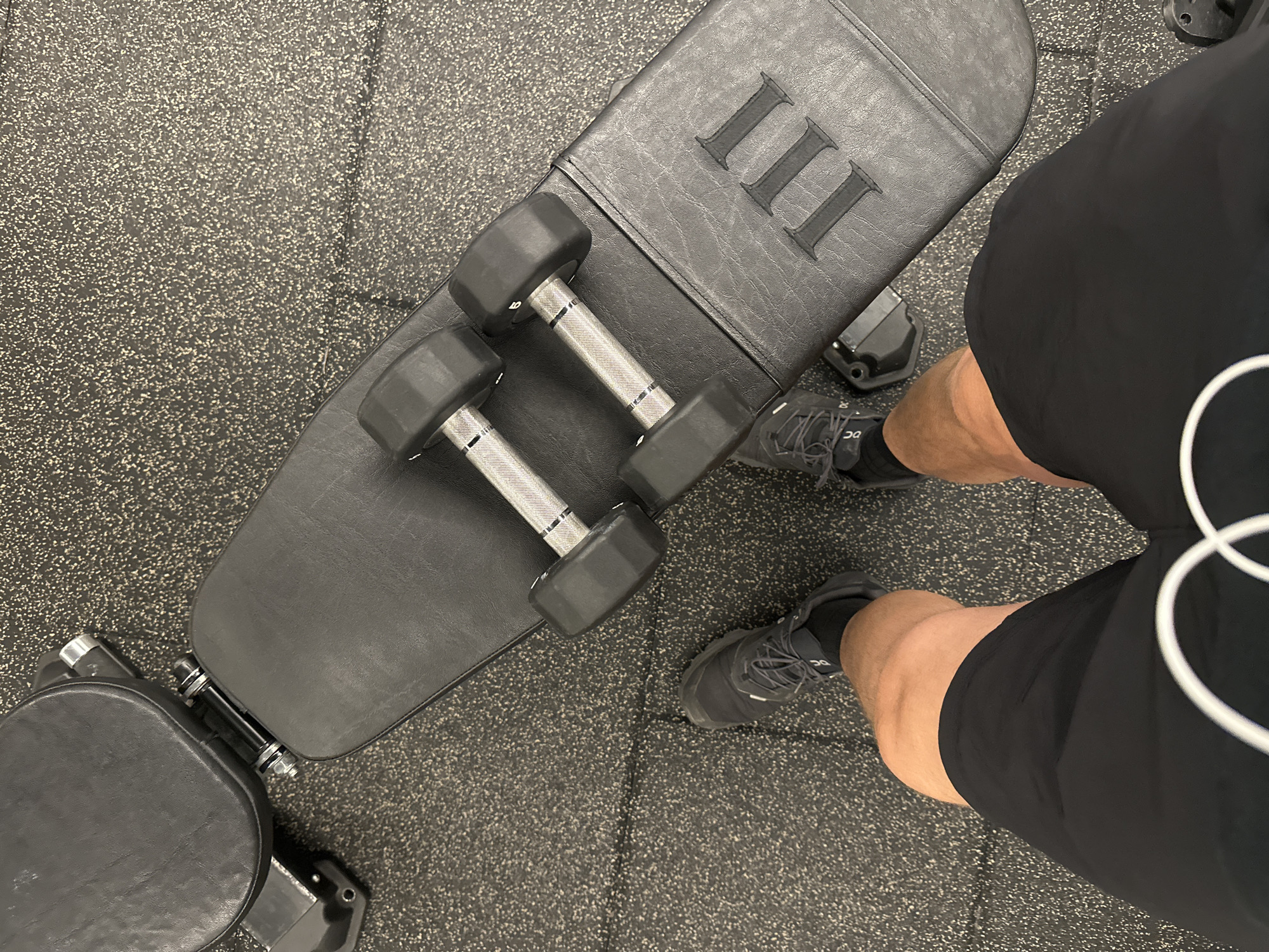 The image shows a high-angle view of a gym setting. A weight bench with a black leather surface and the Roman numeral "III" embossed on it is visible. Two dumbbells rest on the bench. A person wearing black shorts, grey shoes and black socks is standing next to the bench. The floor is covered with a speckled grey rubber mat. The location is not identifiable as London, United Kingdom from the given image.