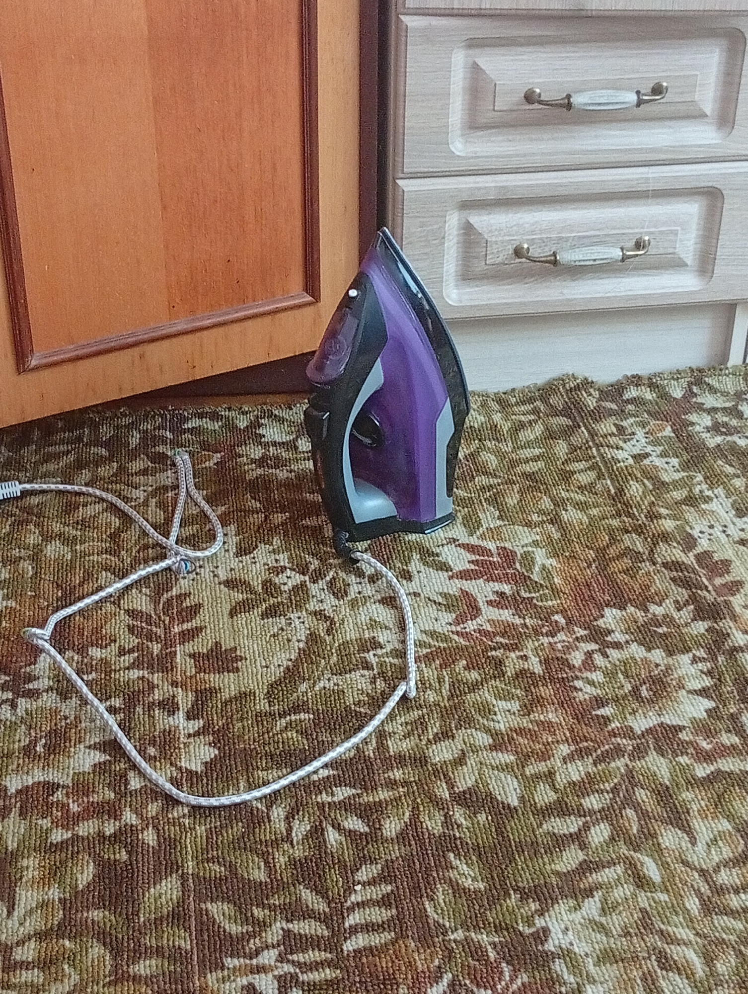 The image shows a purple and black iron with a white cord lying on a floral patterned carpet. To the left of the iron is a wooden cabinet door, and behind it are two drawers with white ceramic and gold handles. The scene is indoors, likely in a domestic setting. There are no people or animals visible in the image. The location is Veliatyno, Ukraine.
