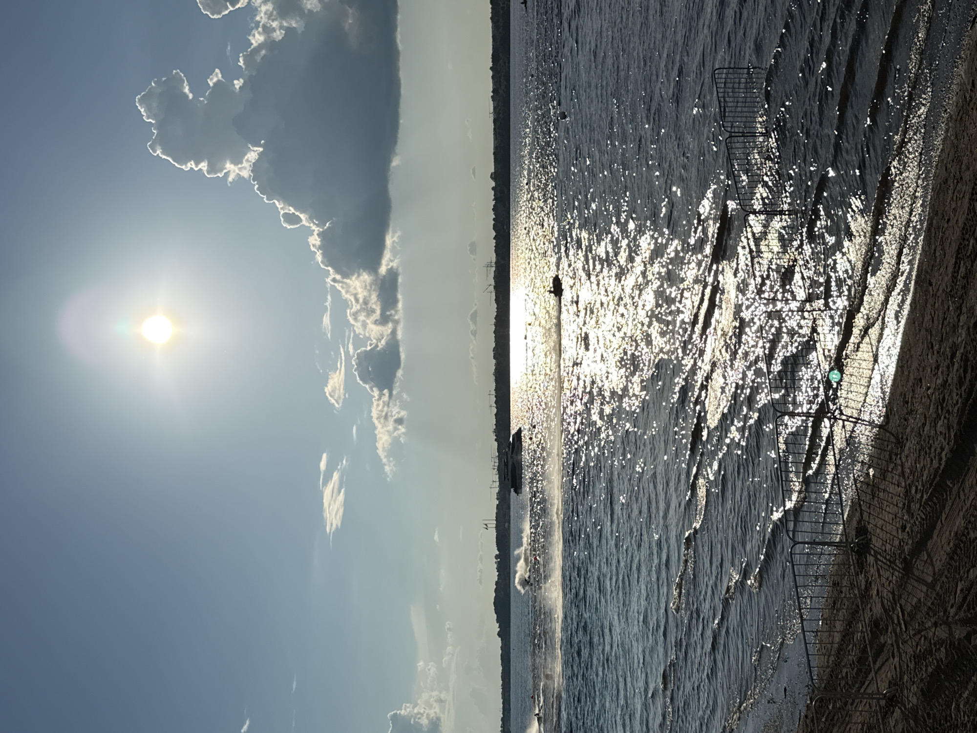 The image captures a scene on a body of water under a bright sun with scattered clouds. The sun is high in the sky, casting a strong glare on the water's surface. In the distance, a large, dark boat is visible, and closer to the foreground, several jet skis are in motion, creating wakes and splashing water. The shoreline in the foreground is sandy with tire tracks, and a low metal fence separates the sand from the water. The water is dark blue with significant sparkle from the sun's reflection. The overall impression is of a recreational activity on a sunny day. The location is the Kama River in the city of Perm, Russia, possibly during a water sports event given the presence of multiple jet skis and buoys.