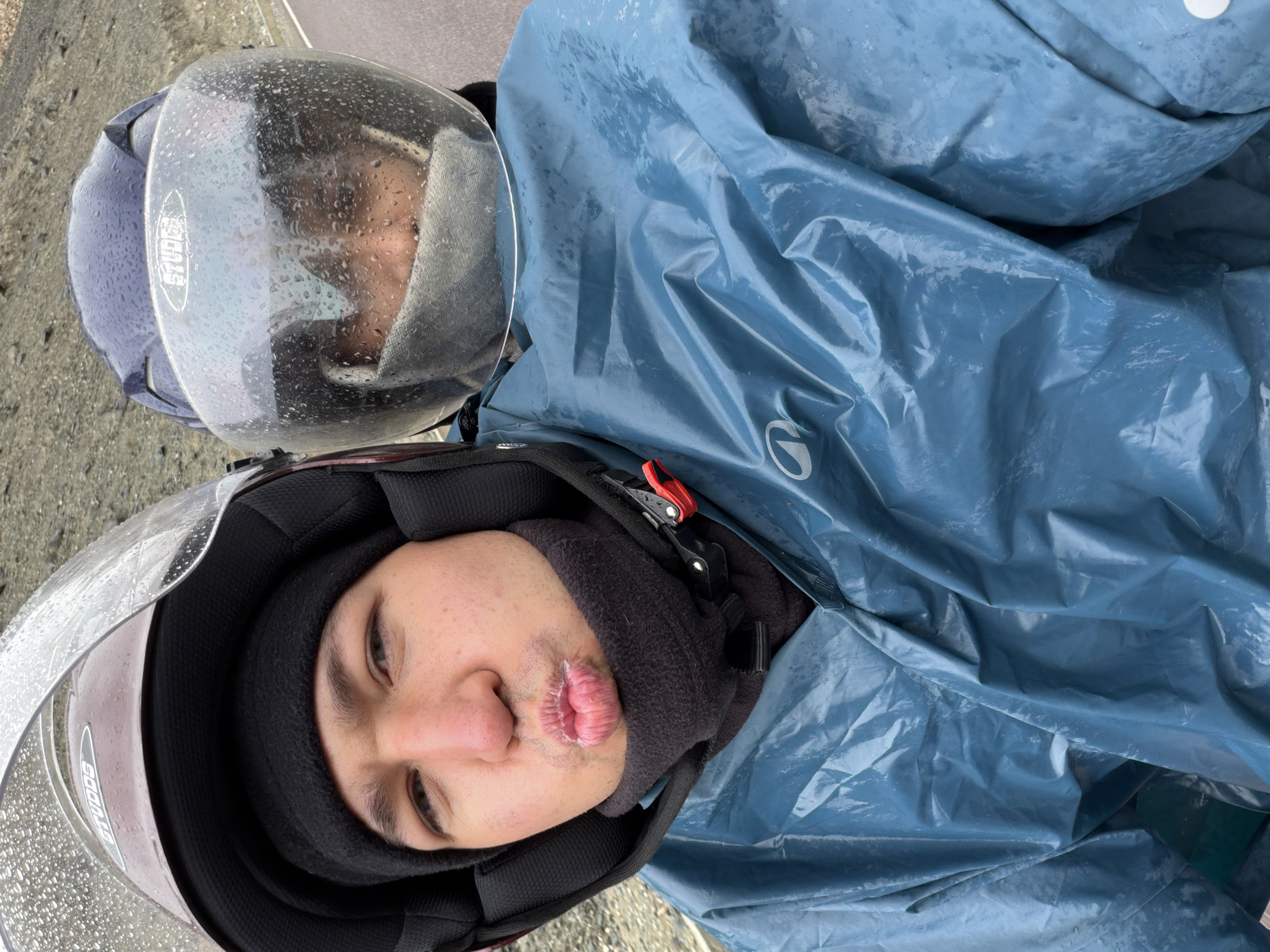 The image shows a close-up view of two individuals on a motorcycle in the city of Sumdo, India. Both individuals are wearing helmets and raincoats, indicating wet weather conditions, likely rain, as evidenced by the water droplets on the helmets' visors and the damp appearance of the raincoats. The person in the foreground, wearing a maroon helmet and a dark neck gaiter, is puckering their lips and looking towards the camera. The second person, behind the first and wearing a dark blue helmet, is partially obscured by the first person's helmet and is wearing a light-colored face mask. The background suggests a mountainous or hilly terrain, with a road visible. The overall scene depicts a moment captured during a motorcycle ride in inclement weather in Sumdo.