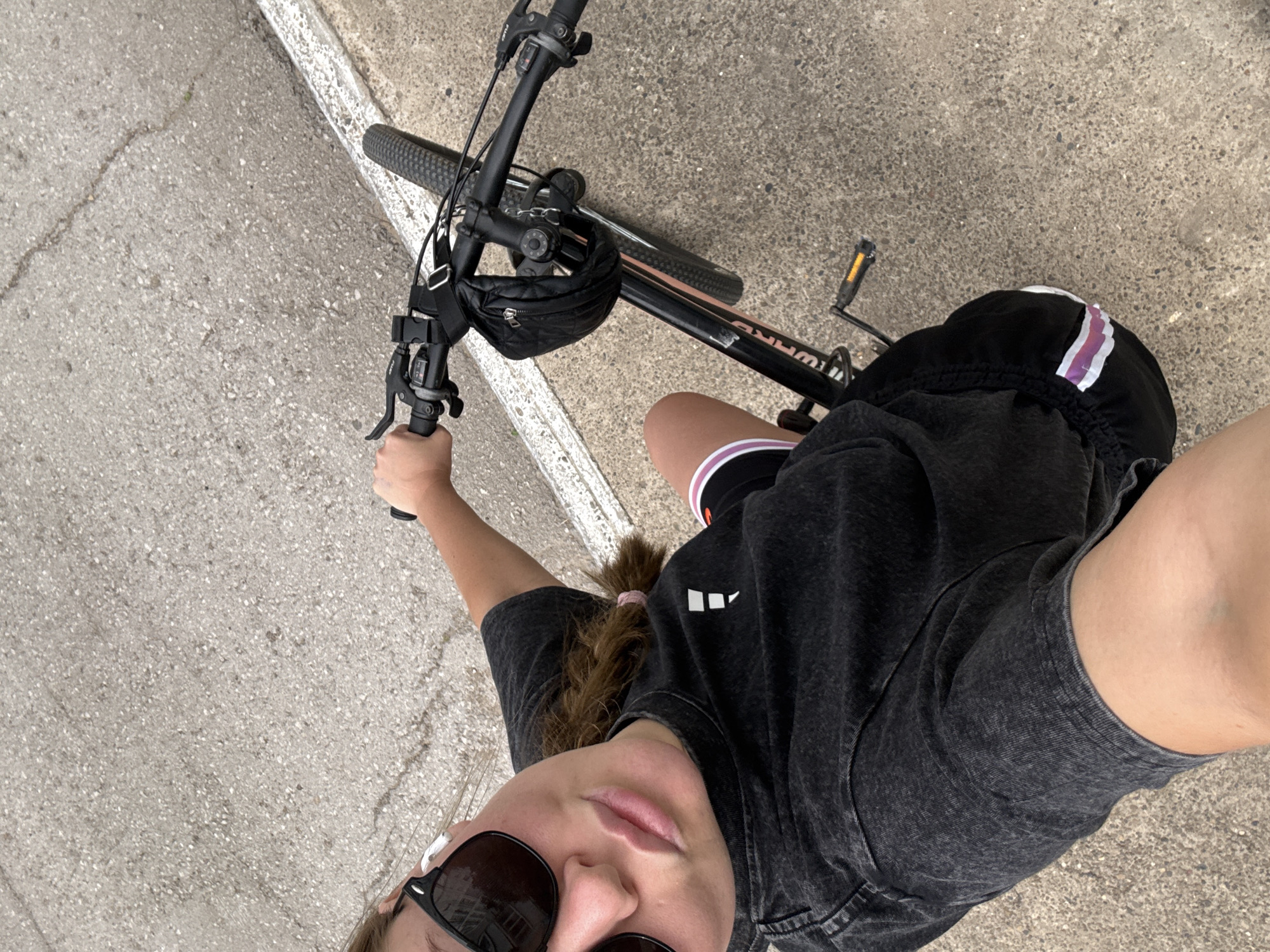 An overhead, first-person perspective shot shows a person on a black mountain bike on a paved surface. The person is wearing black sunglasses, white earbuds, and a black t-shirt. Their left hand is on the handlebars, and their right arm is extended toward the camera. The bicycle has black handlebars with a black pouch attached, black grips, and a black frame with pink accents. The paved surface is gray with visible cracks and has a white line along the right edge. The location is identified as Chaikovsky, Russia.