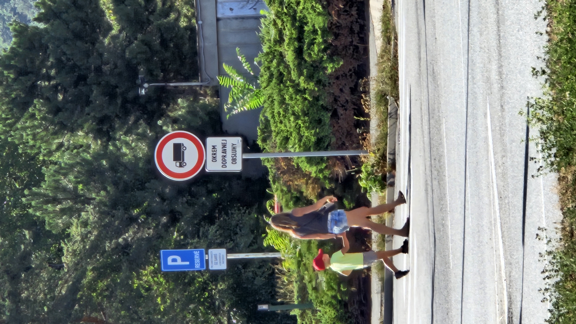 In Prešov, Slovakia, a woman and a young boy are crossing a street. The woman, wearing denim shorts and a dark tank top, is holding the boy's hand. The boy is dressed in a light green t-shirt and grey shorts, with a red cap on his head. They are walking on a pedestrian crossing, indicated by white lines on the asphalt. In the background, there are signs. One is a blue parking sign with a "P" and the word "RESERVÉ." Another is a circular red and white sign with a black truck silhouette, indicating a restriction for trucks. Below this sign, a rectangular white sign displays text in Slovak. Lush green bushes and trees line the street, with a building visible further in the background. The scene appears to be captured on a bright, sunny day.
