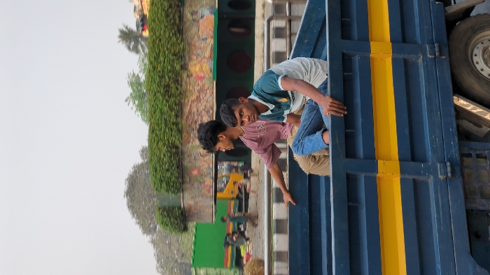 The image displays two young males riding in the open cargo bed of a dark blue truck featuring a yellow horizontal stripe, observed within an urban environment in Dhaka, Bangladesh. The male on the left, wearing a short-sleeved reddish-brown shirt and light-colored trousers, looks to his left. The male on the right, dressed in a green, white, and blue striped polo shirt and blue jeans, looks directly at the viewer. Both individuals are seated with their hands resting on the truck's upper side rails. In the background, a multi-lane road is visible with several vehicles, including a prominent green container truck and a yellow truck, alongside blurred figures of pedestrians. An elevated concrete structure, densely covered with green foliage, and a wall adorned with multi-colored graffiti are also present. The sky is light gray and appears hazy.