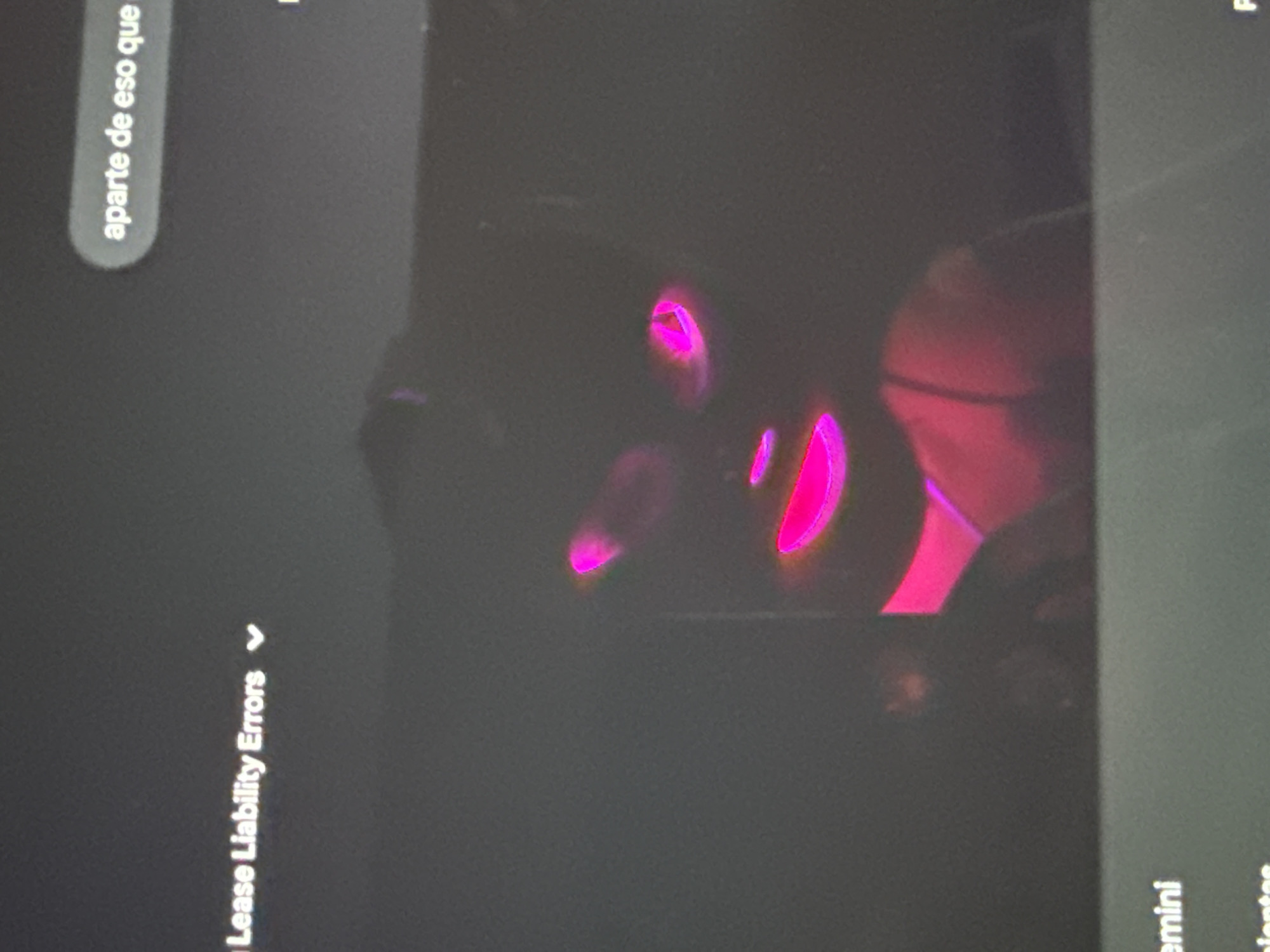 The image displays a dark scene, primarily focusing on a digital screen or display in a dimly lit environment. The central visual elements are several bright, glowing shapes in vibrant pink and magenta, which appear to be reflections or illuminated components. These luminous shapes create an abstract pattern against the dark background and could potentially be highlights on a person's face, glasses, or reflective elements of an object.
Visible text indicates the presence of a screen interface. In the top left corner, a rounded gray box contains the partially visible Spanish phrase "aparte de eso que" (meaning "besides that"). Towards the bottom left, the text "...Lease Liability Errors" is discernible, accompanied by a dropdown arrow, suggesting content related to business, finance, or a system interface. On the far right, in a slightly lighter gray section, fragments of words such as "emini" and "ientes" are visible.
The overall lighting is extremely low, suggesting the photograph was taken at night or in a very dark room. The sharp contrast of the glowing pink/magenta against the dark gray and black tones is a prominent feature. The scene offers no specific visual cues to its location in San Bernardino Tlaxcalancingo, Mexico, other than the provided context. No specific activity or emotions are explicitly visible, beyond the implied interaction with a digital display in a dark setting.