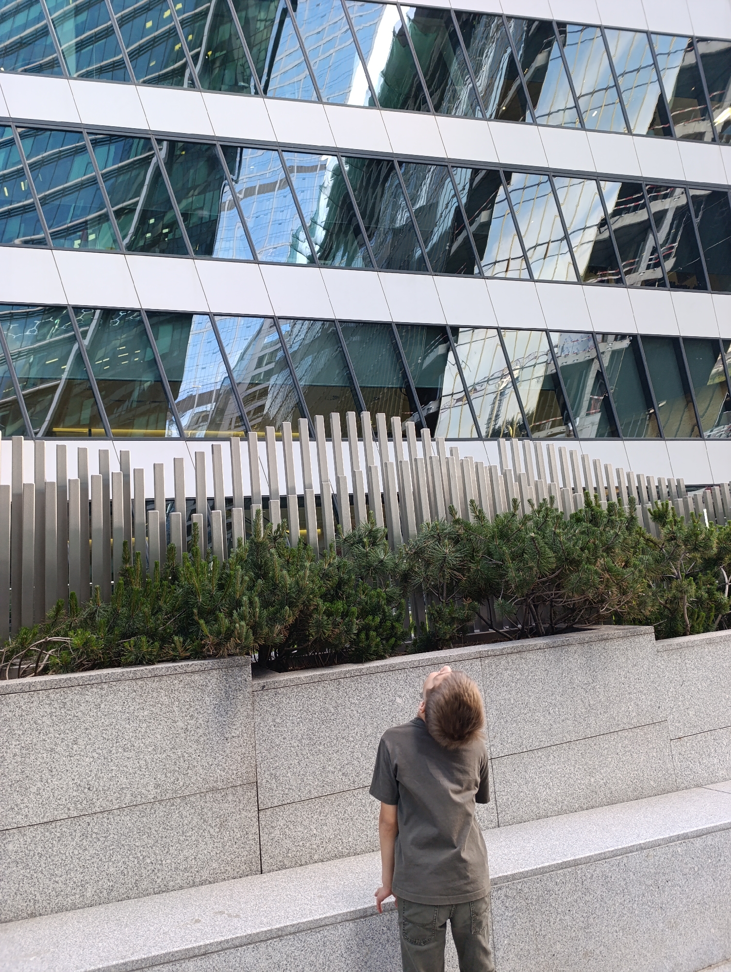 A person, appearing to be a young boy or teenager, stands with their back to the camera, looking upwards at a modern skyscraper. They are dressed in a dark grey t-shirt and greyish-green pants. The setting is an urban outdoor area, likely a plaza or sidewalk in Moscow, Russia.
In the foreground and midground, there is a multi-tiered structure made of light grey, speckled granite blocks, serving as both a wall and a low bench. Behind this structure, lush green coniferous shrubs are planted in a raised bed. Further back, a decorative fence or barrier is visible, composed of numerous vertical, irregularly spaced, light grey posts of varying heights.
Dominating the background is a tall, contemporary building with a striking glass facade. The building features alternating horizontal bands of white material and sections of dark, reflective glass. These glass sections are divided into a repeating pattern of diagonal panels, creating a dynamic, faceted appearance. The glass reflects the sky and other nearby structures, including parts of what appears to be another building and a bright, possibly cloudy, sky.
The scene is illuminated by natural daylight, suggesting it is daytime with clear or partly cloudy weather. The person's upward gaze implies a sense of awe or curiosity towards the impressive architecture. No discernible text is visible in the image.