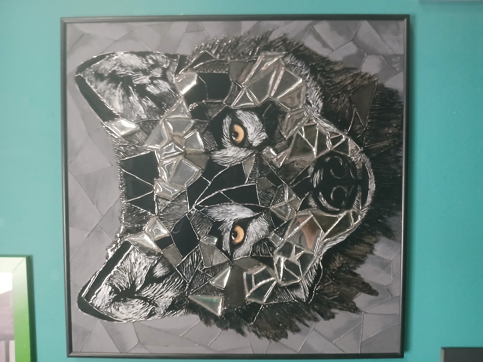 The image shows a framed artwork of a wolf's face, made from what appears to be shattered glass or mirror pieces arranged in a mosaic style. The wolf's eyes are a prominent amber color, and the fur is depicted with black, white, and gray tones. The background of the artwork is a muted gray, with a geometric pattern. The artwork is framed in a dark material and hangs on a teal-colored wall. There is another framed artwork with a green border visible in the upper left corner, partially out of focus. The image is taken from a medium distance, with the artwork as the central subject.