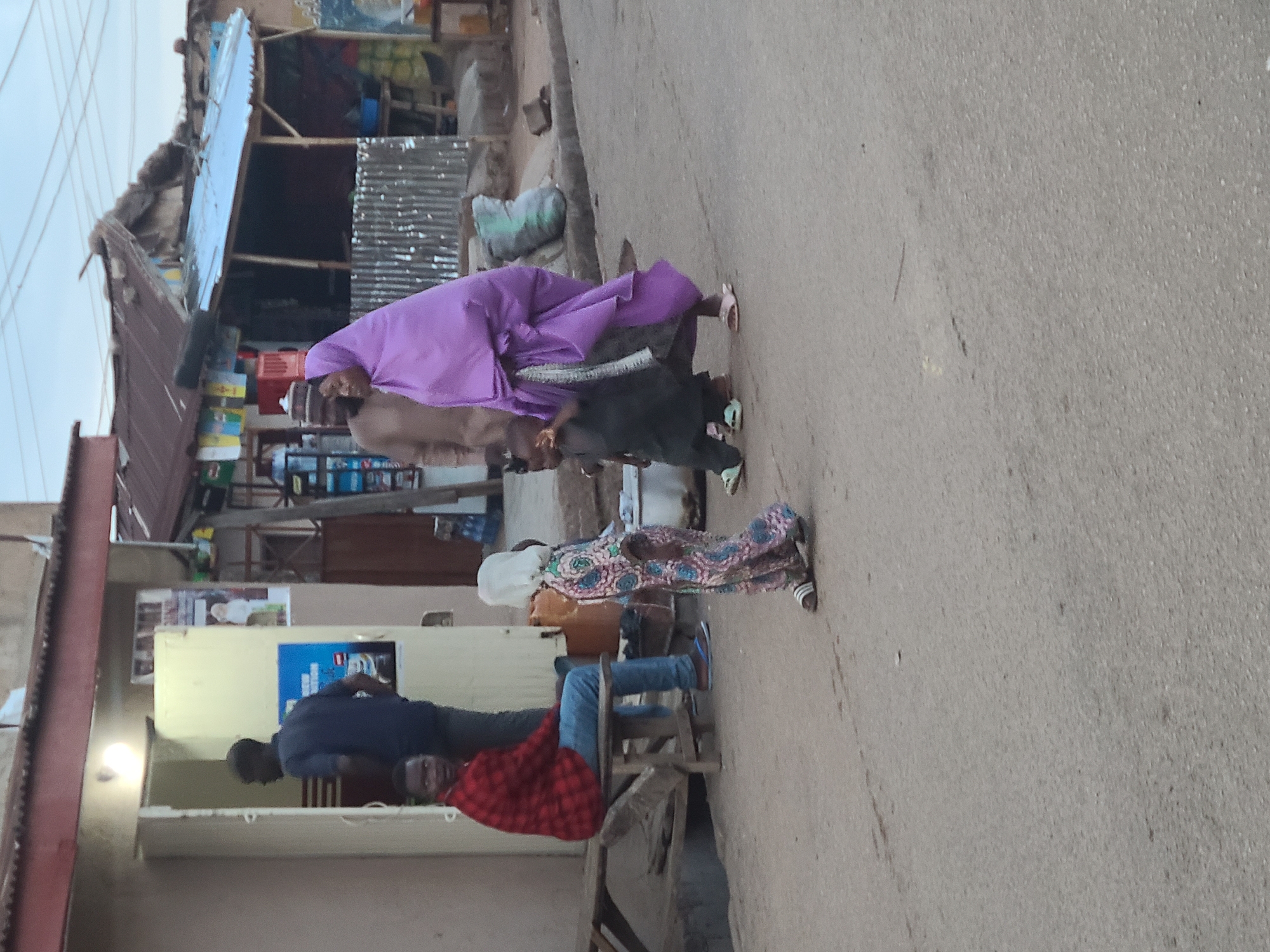 The image depicts an outdoor street scene, likely in a bustling town or market, under an overcast sky or during the late afternoon/early evening. The perspective is tilted, showing an unpaved or lightly graveled road surface on the right and various structures on the left.
Multiple individuals are present. In the mid-ground, a person wearing a vibrant purple head covering and loose garment stands alongside another person in dark clothing, possibly a child. Behind them, a person in a light-colored, traditional garment is partially visible. Further left, a person in a patterned long dress stands. In the foreground, two men are seated on simple wooden stools; one wears a dark blue shirt and jeans, and the other, facing the viewer, is in a red plaid shirt.
The background is lined with rudimentary structures, appearing to be small shops or stalls, constructed from corrugated metal sheets and wooden frames. These structures display various items, including stacked containers and a large green sack. An electrical wire runs overhead against the pale sky. A single bright light bulb illuminates a wall section where several posters or signs are affixed, though any text on them is indistinct and illegible. The scene conveys a sense of everyday activity and street-side commerce.