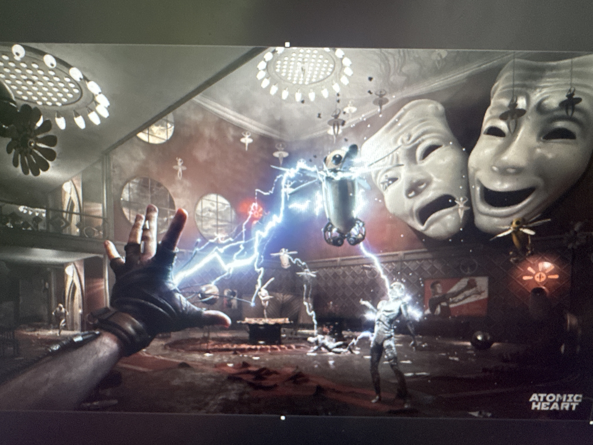 The image depicts a scene from the video game "Atomic Heart." In the foreground, a gloved hand reaches out towards a chaotic environment. The setting appears to be an interior space with ornate architecture, including a large chandelier on the ceiling and decorative circular windows. Dominating the background are two large theatrical masks, one depicting a tragic expression and the other a comedic one, both adorned with small dangling figures resembling ballerinas.
The central action involves a robot-like figure standing amidst an electrical storm, with bolts of blue lightning emanating from its position. Other robotic figures and what appear to be fallen individuals are scattered on the ground, suggesting a conflict. Various metallic objects and debris are present throughout the scene. A poster with a stylized image of a person can be seen on the wall. The overall atmosphere is one of disarray and technological combat. The game's title, "ATOMIC HEART," is visible in the bottom right corner. There is no discernible connection to the city of Stepnogorsk or its city administration in Kazakhstan within this image.