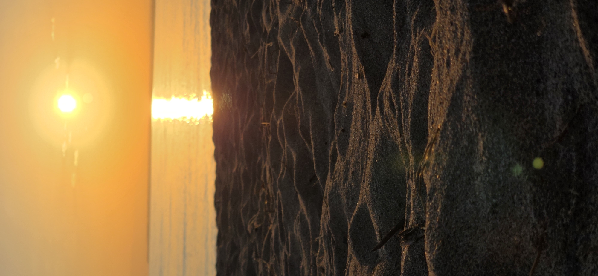 The image is a vertical, low-angle shot of a beach at sunset. The foreground is dominated by textured sand dunes, with small pieces of debris scattered across the surface. In the background, the sun is setting over a calm body of water, casting a bright reflection on the surface. The sky is a warm orange hue, typical of a sunset. The scene is devoid of any people or specific landmarks that would indicate a location within Basbeshik, Kyrgyzstan.