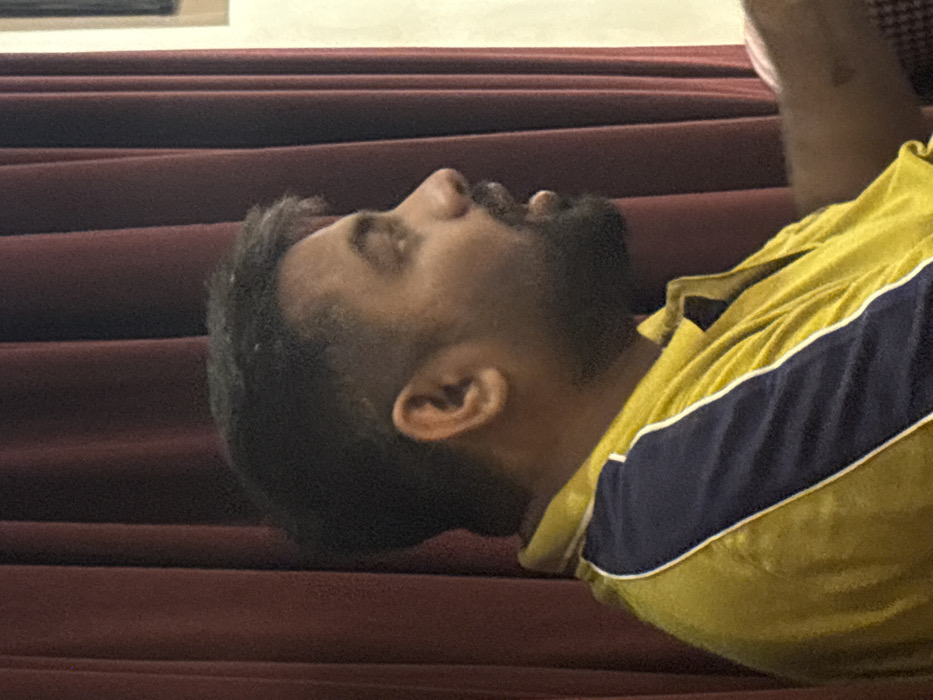 An indoor, medium-close shot captures a male subject in right profile. The subject exhibits a medium-dark complexion, short dark hair with a faded cut, and a full dark beard and mustache. His mouth is slightly open. He wears a yellow polo shirt with a dark blue panel outlined by a white stripe on the left shoulder/upper arm area. The background features dark red, vertically pleated curtains to the left and a lighter, cream-toned wall section to the right. A faint, dark mark is present on the subject's visible right forearm. The image provides no visual indicators to confirm the specified location of Howrah, India.